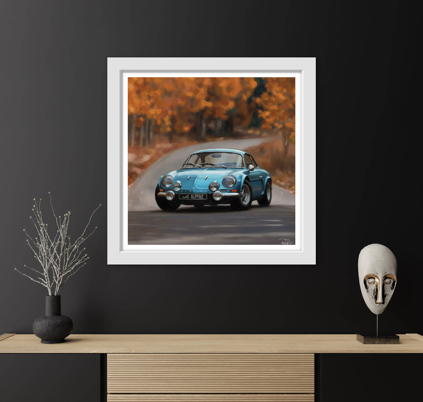 Framed artwork of a blue vintage car on a road with autumn trees, displayed on a dark wall above a wooden cabinet.
