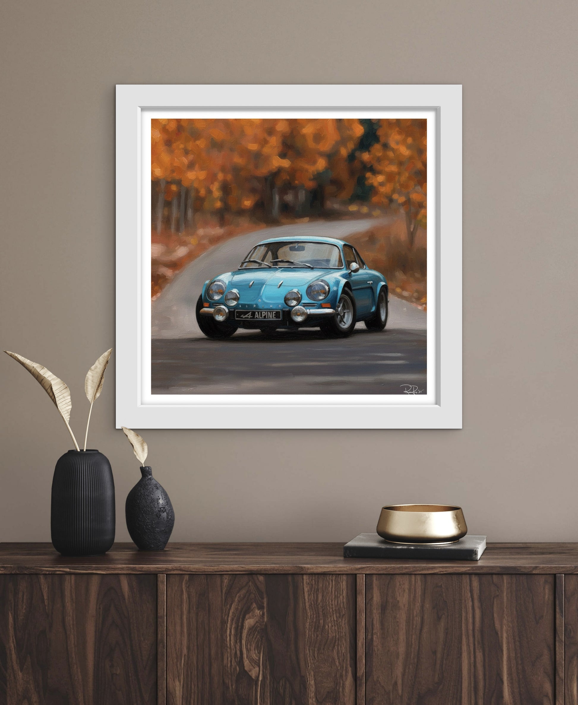 Framed artwork of a blue car on a road with autumn trees, displayed on a wooden shelf.