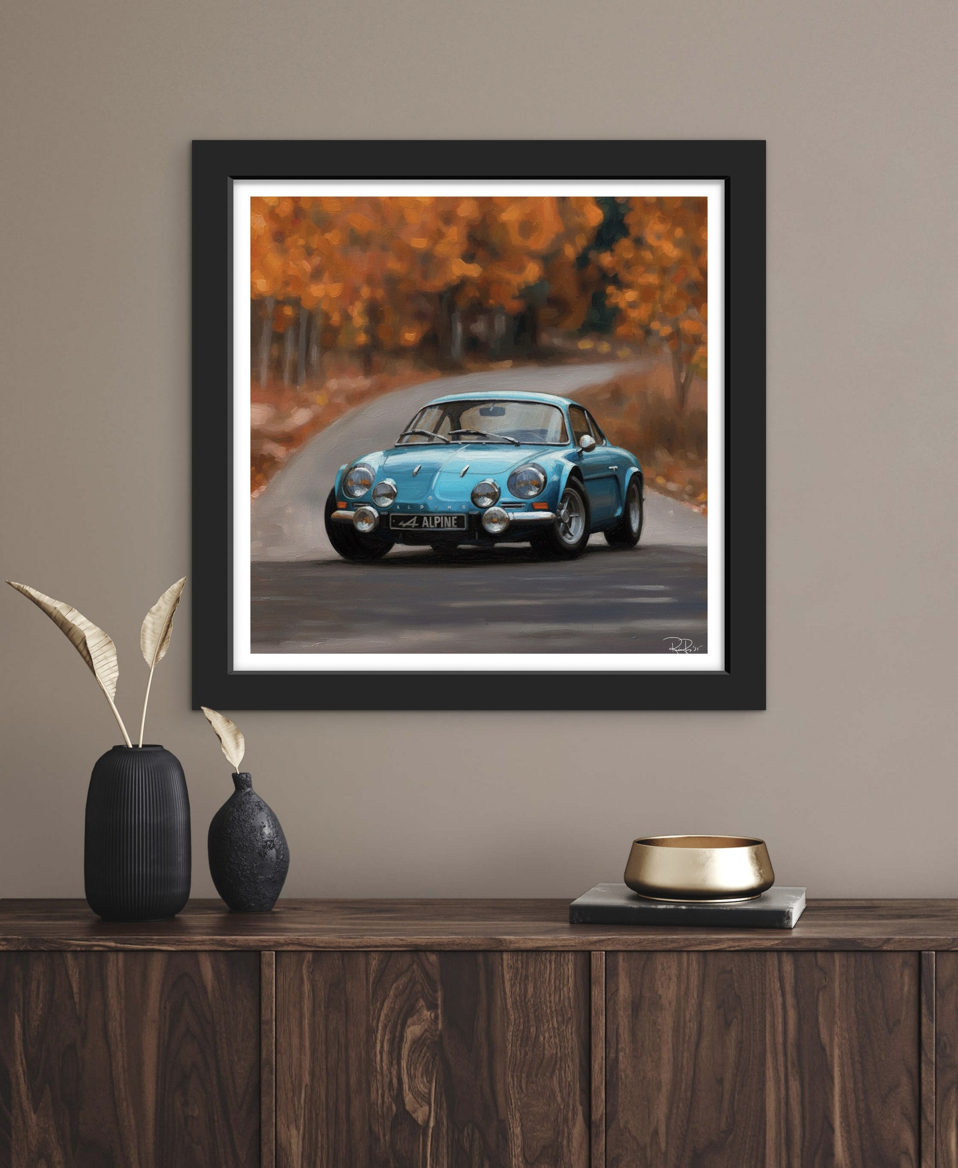 Framed artwork of a blue car on a road with autumn trees, displayed on a wall above a wooden console table.