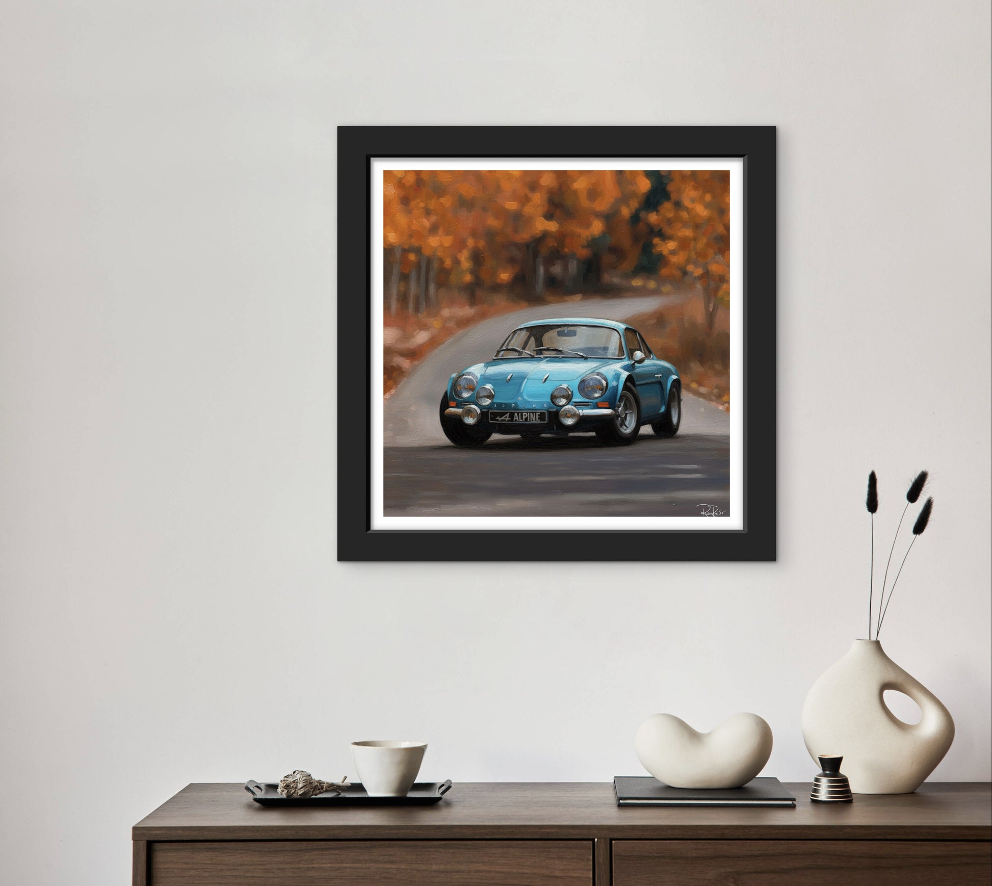 Framed photograph of a blue car on a road with autumn trees, displayed on a wall above a wooden console table.