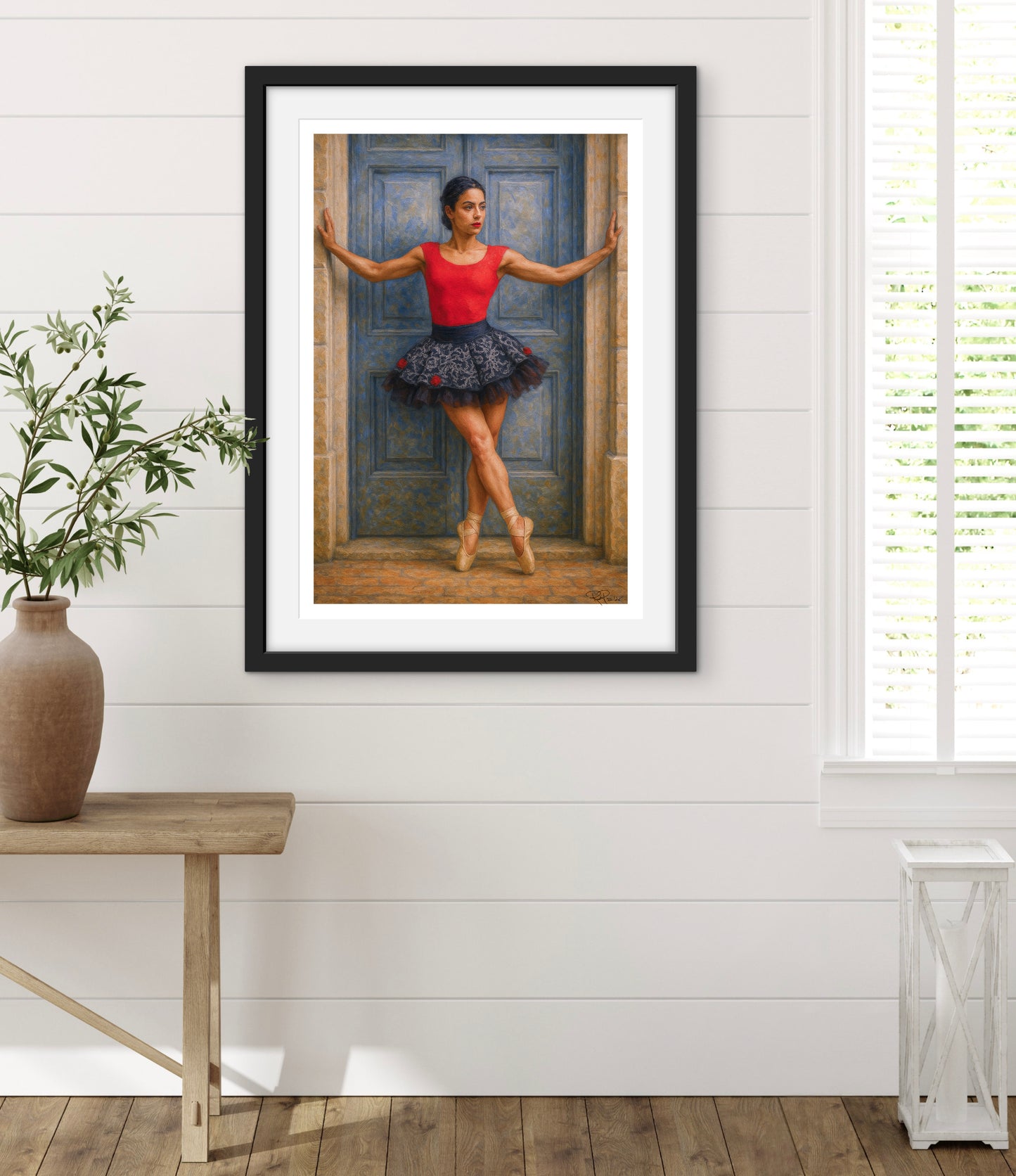 Framed artwork of a ballerina in a red top and floral skirt against a blue door, displayed in a room with a wooden floor and white walls.
