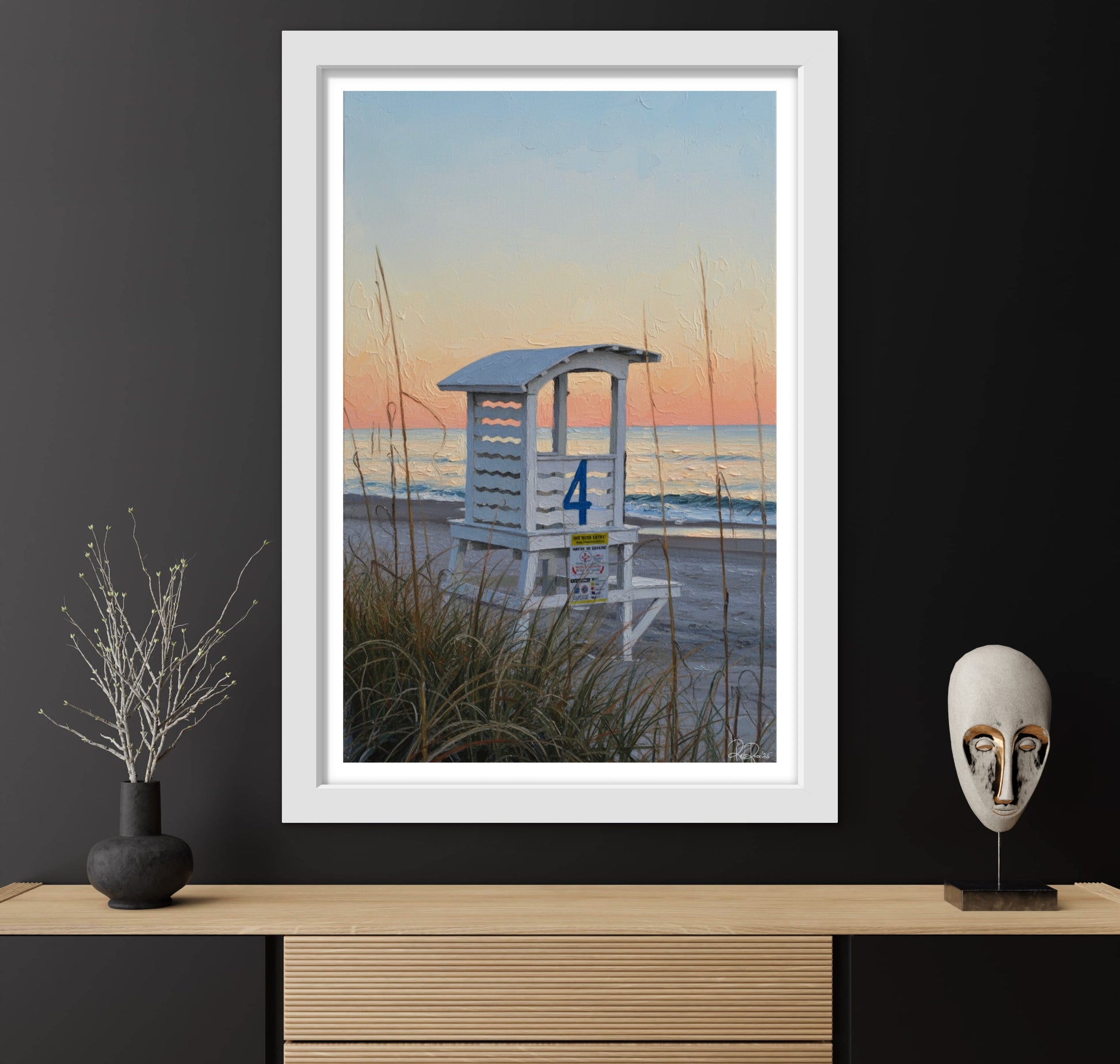 Framed artwork of a lifeguard stand on a beach with a white mask and plant on a wooden surface.