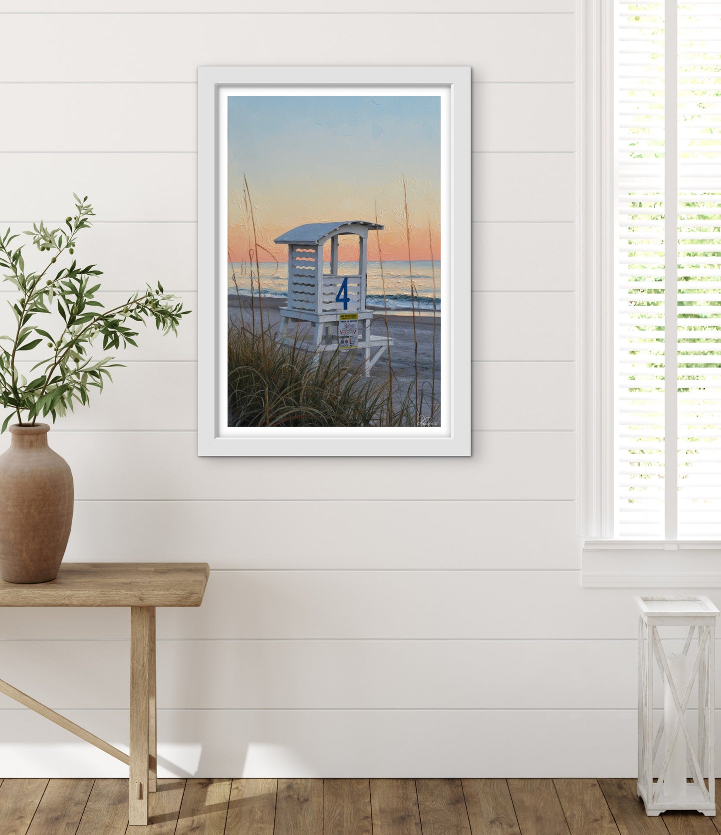 Framed beach scene print on a wall with a wooden table and plant in the foreground.