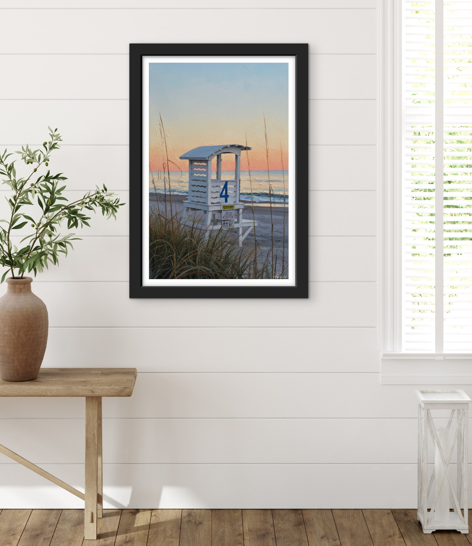 Framed artwork of a beach scene with a lifeguard stand on a white wall, next to a wooden table with a plant.