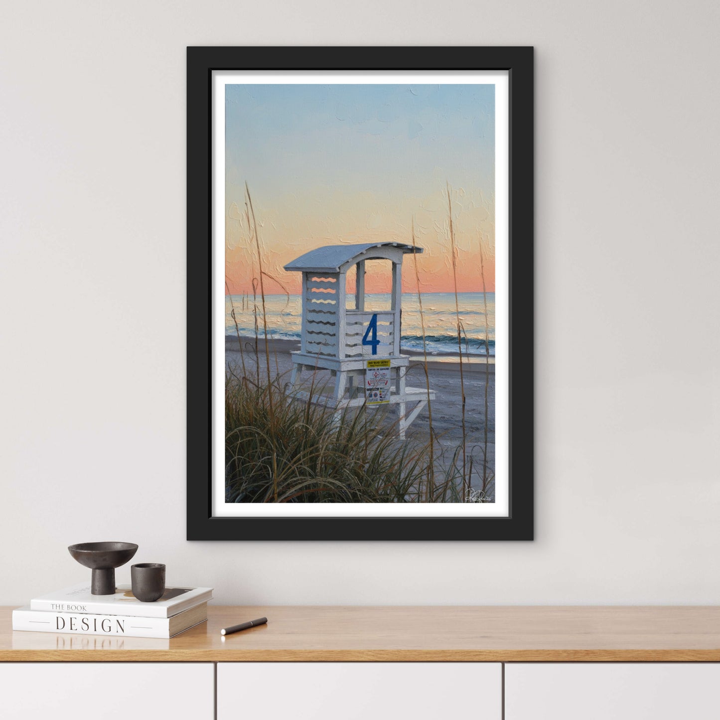 Framed artwork of a beach scene with a lifeguard tower on a wall above a wooden surface.