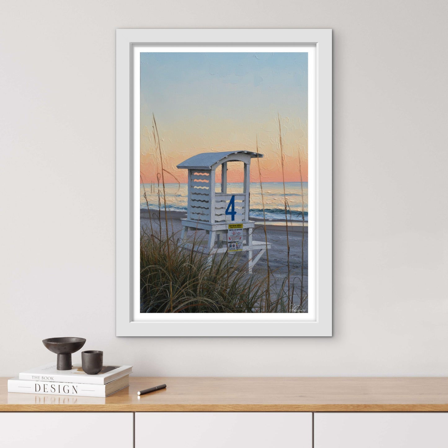 Framed artwork of a beach scene with a lifeguard stand on a wall above a wooden surface.