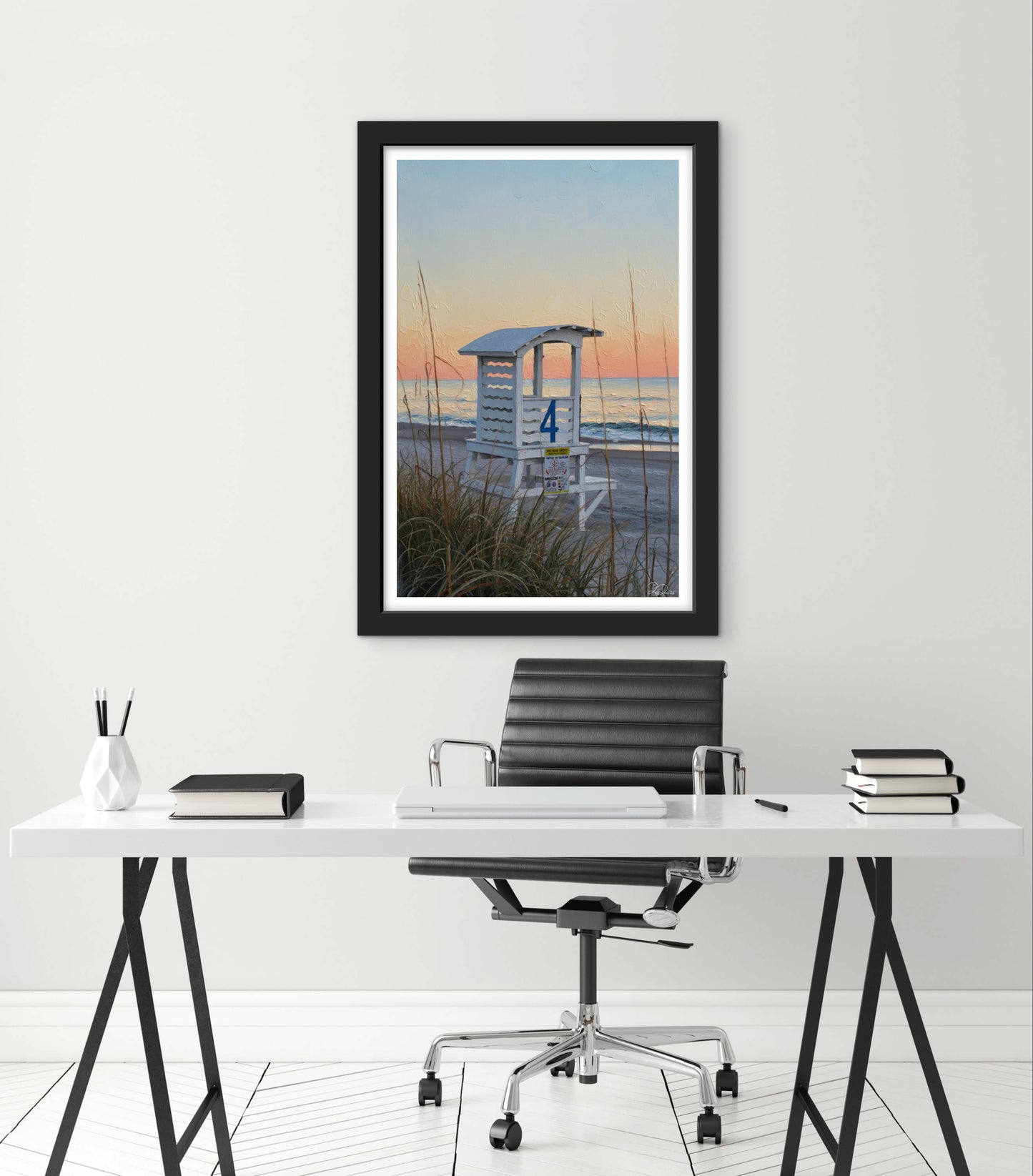 Framed artwork of a lifeguard tower over water on a white wall above a desk.
