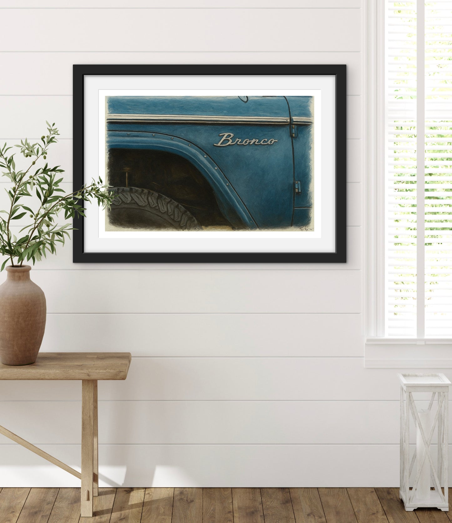 Framed picture of a blue Ford Bronco on a white wall with a wooden bench and plant.