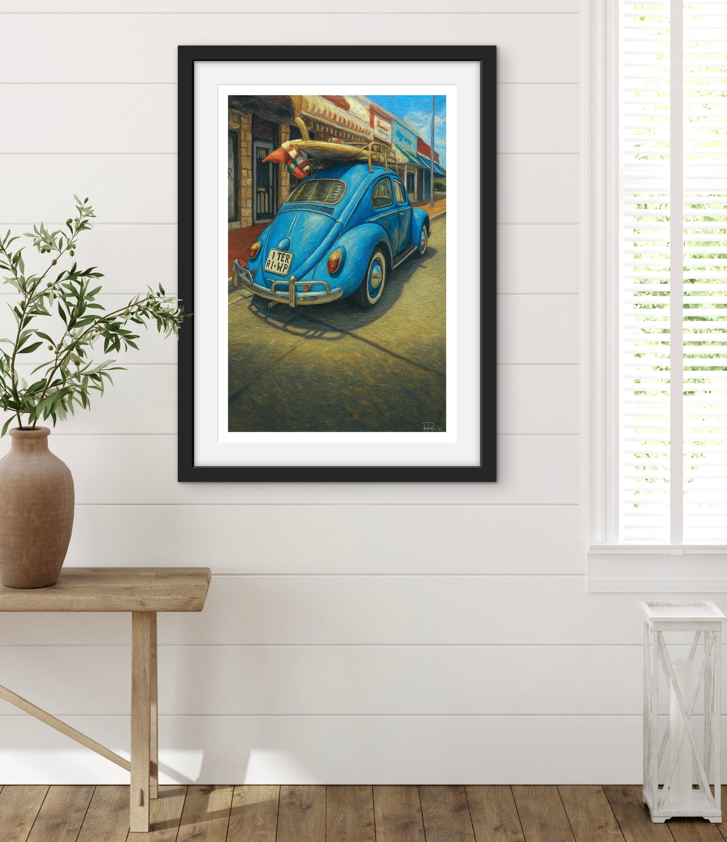 Framed artwork of a blue vintage car on a white wall with a wooden floor and plant.