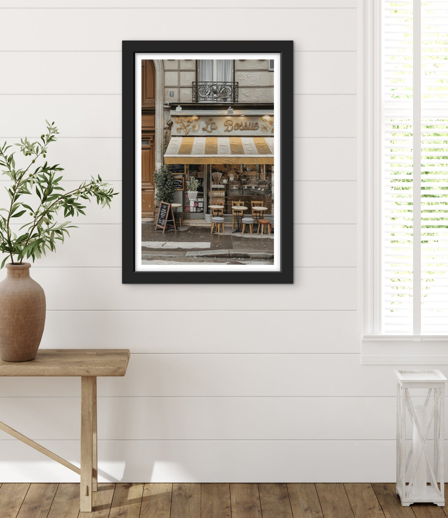 Framed photograph of a street scene with a store front on a white wall, next to a wooden table with a plant.