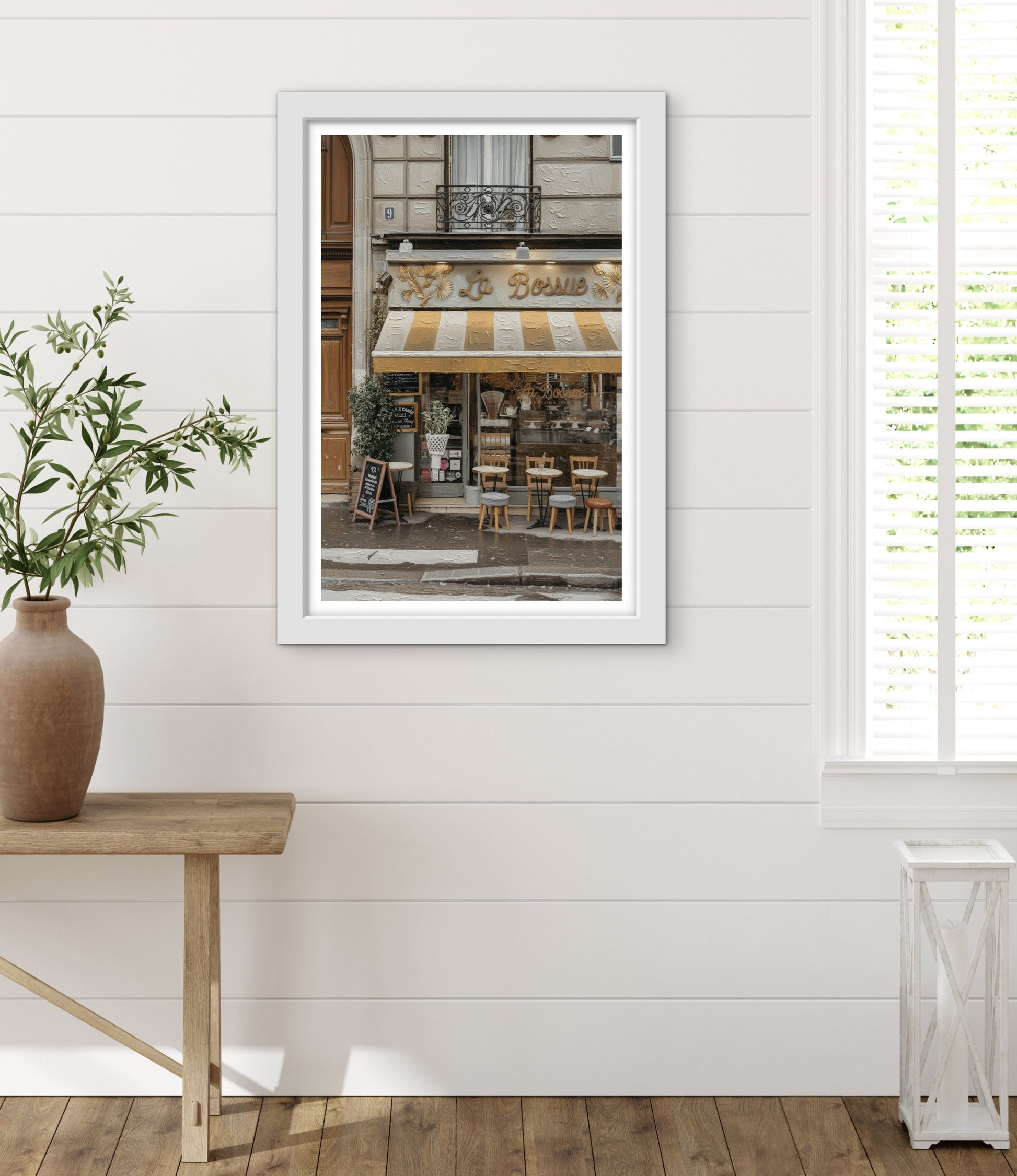 Framed photograph of a cafe on a white wall with a wooden table and plant in the foreground.
