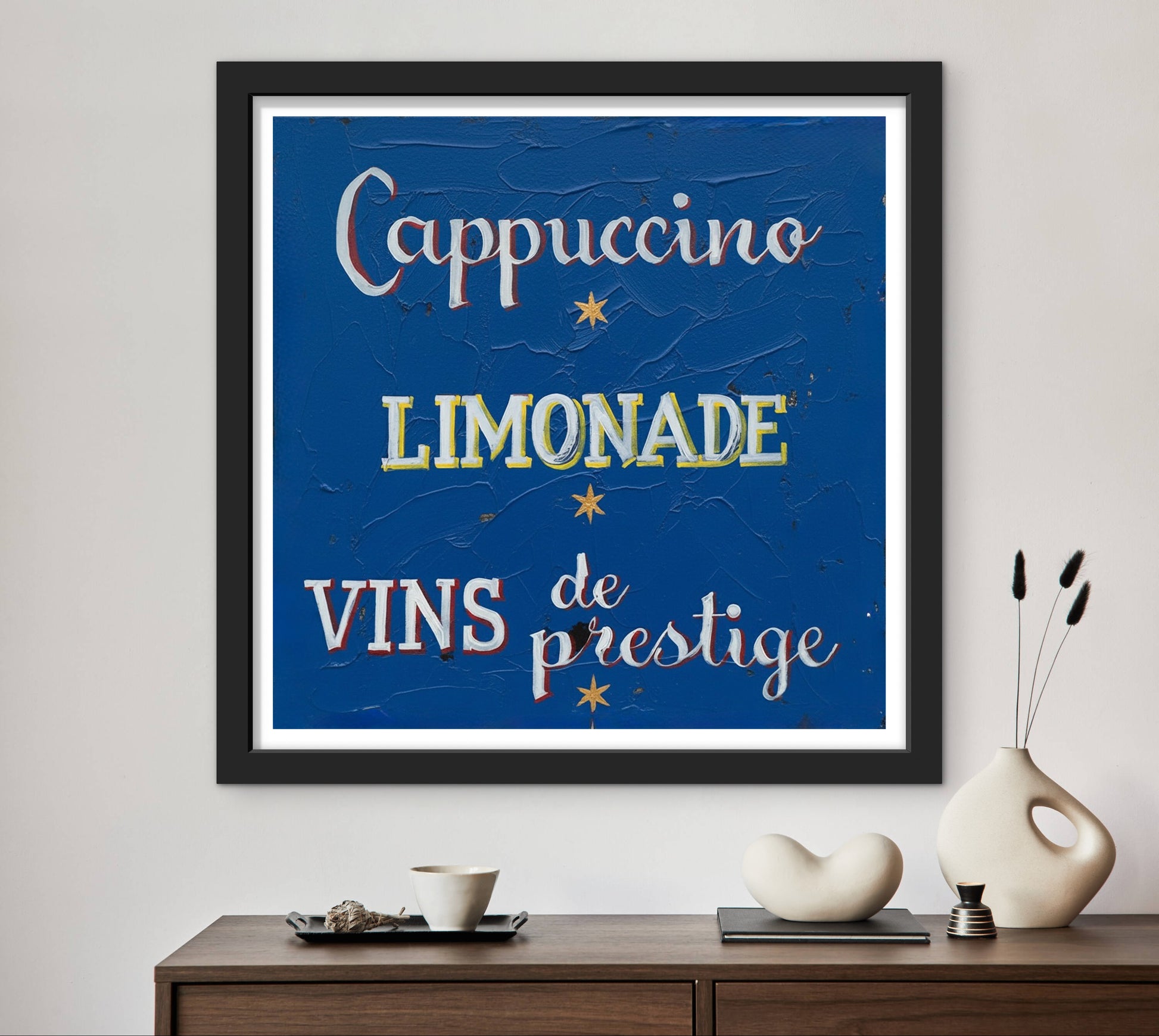 Framed artwork with text 'Cappuccino', 'Limonade', 'Vins de prestige' on a blue background, displayed on a wall above a wooden console table.