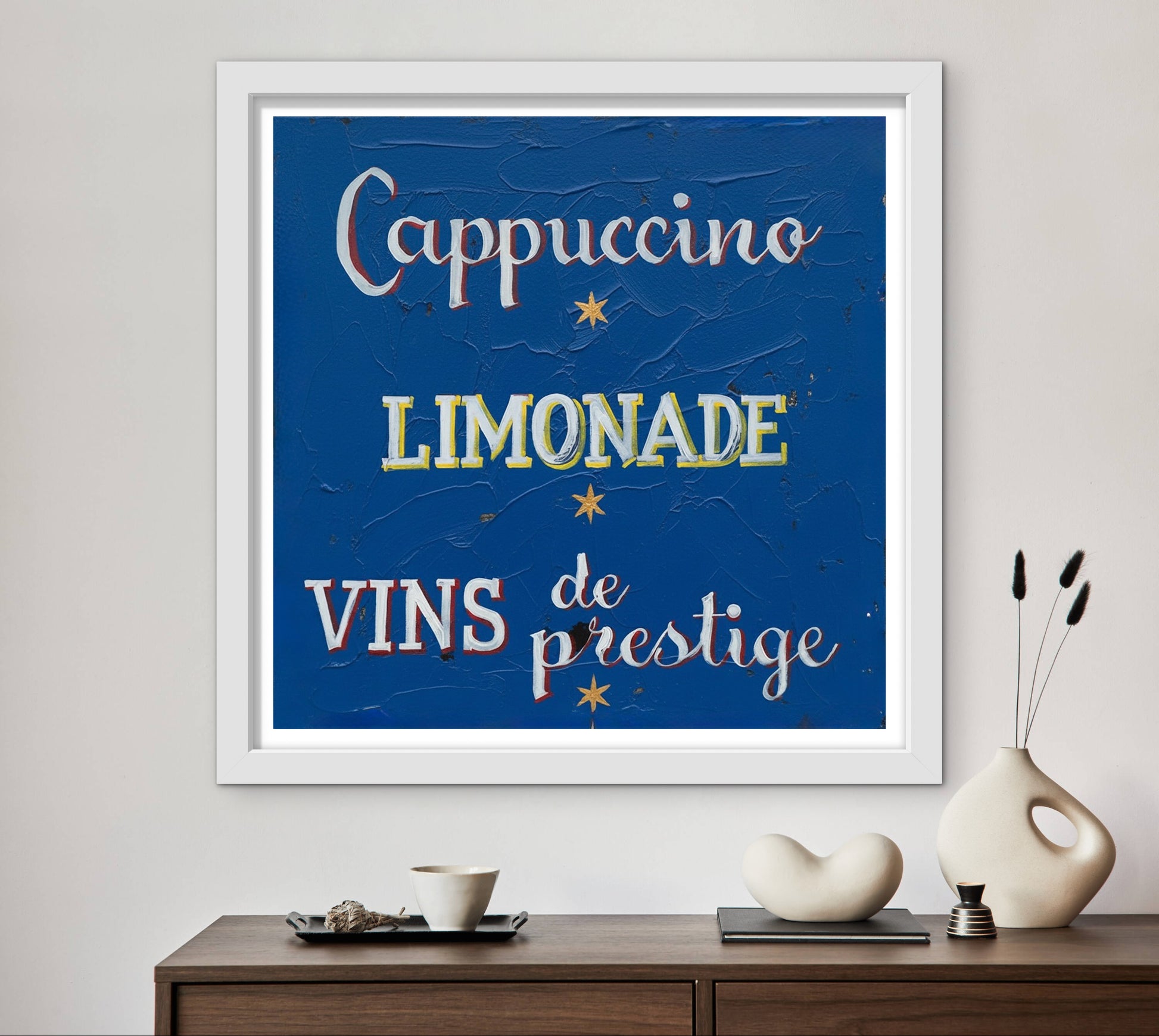 Framed artwork with 'Cappuccino', 'Limonade', and 'Vins de prestige' text on a blue background, displayed on a wall above a wooden console table.