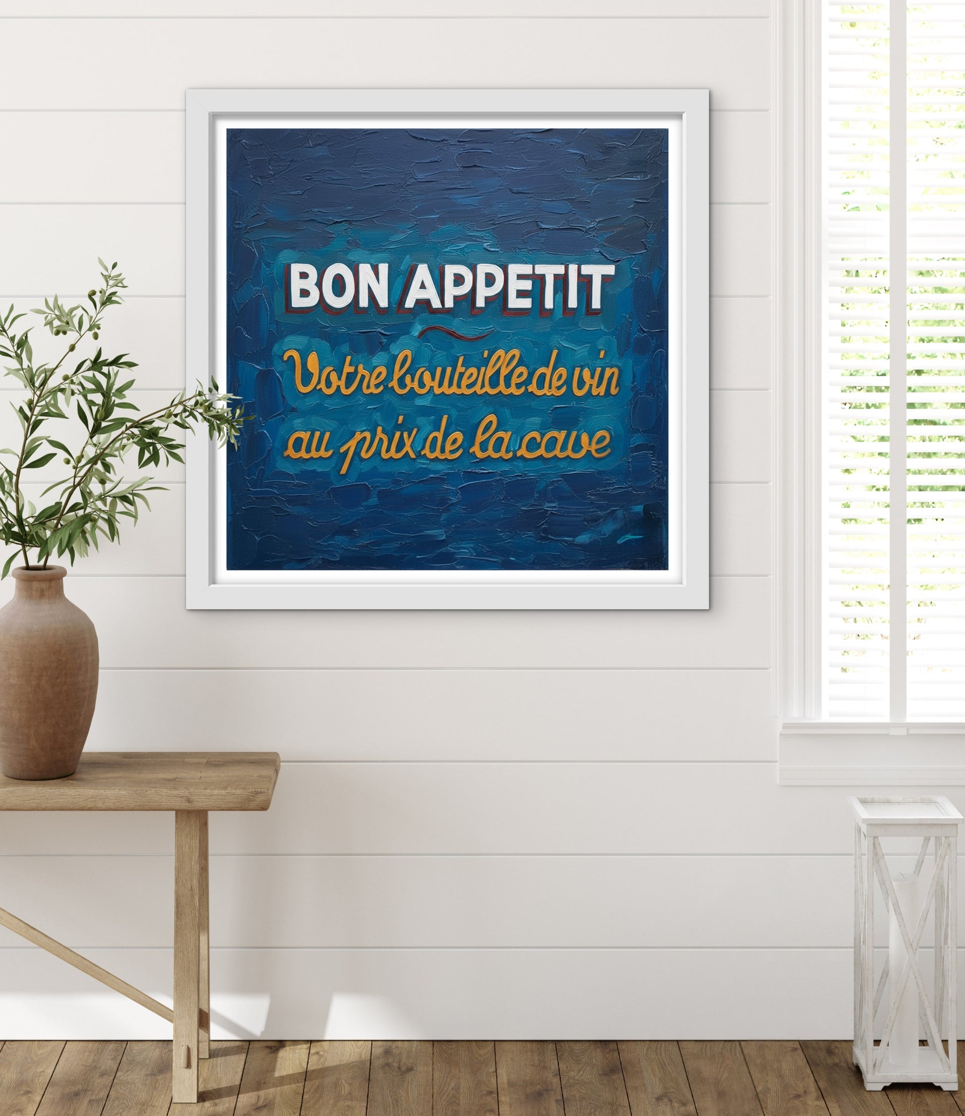Framed artwork with 'Bon Appetit' text on a blue background in a room with a plant and wooden table.