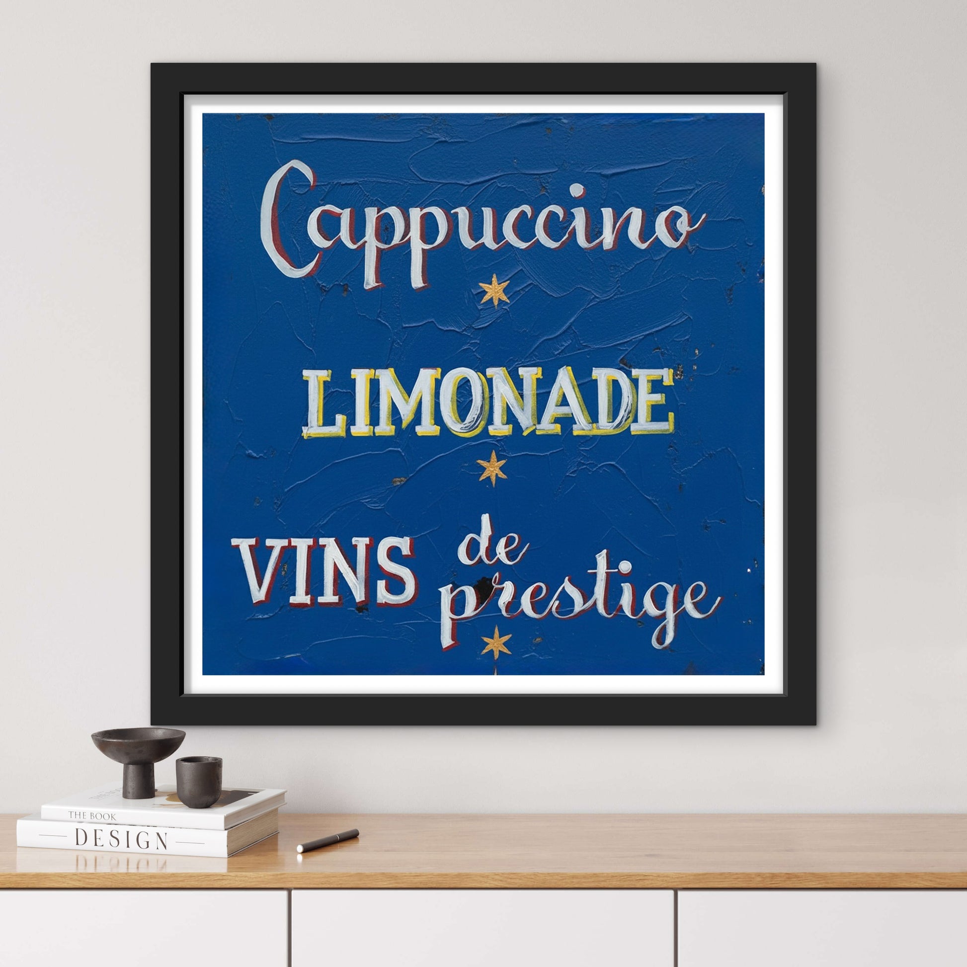 Framed artwork with text 'Cappuccino', 'Limonade', 'Vins de prestige' on a blue background, displayed on a wall.