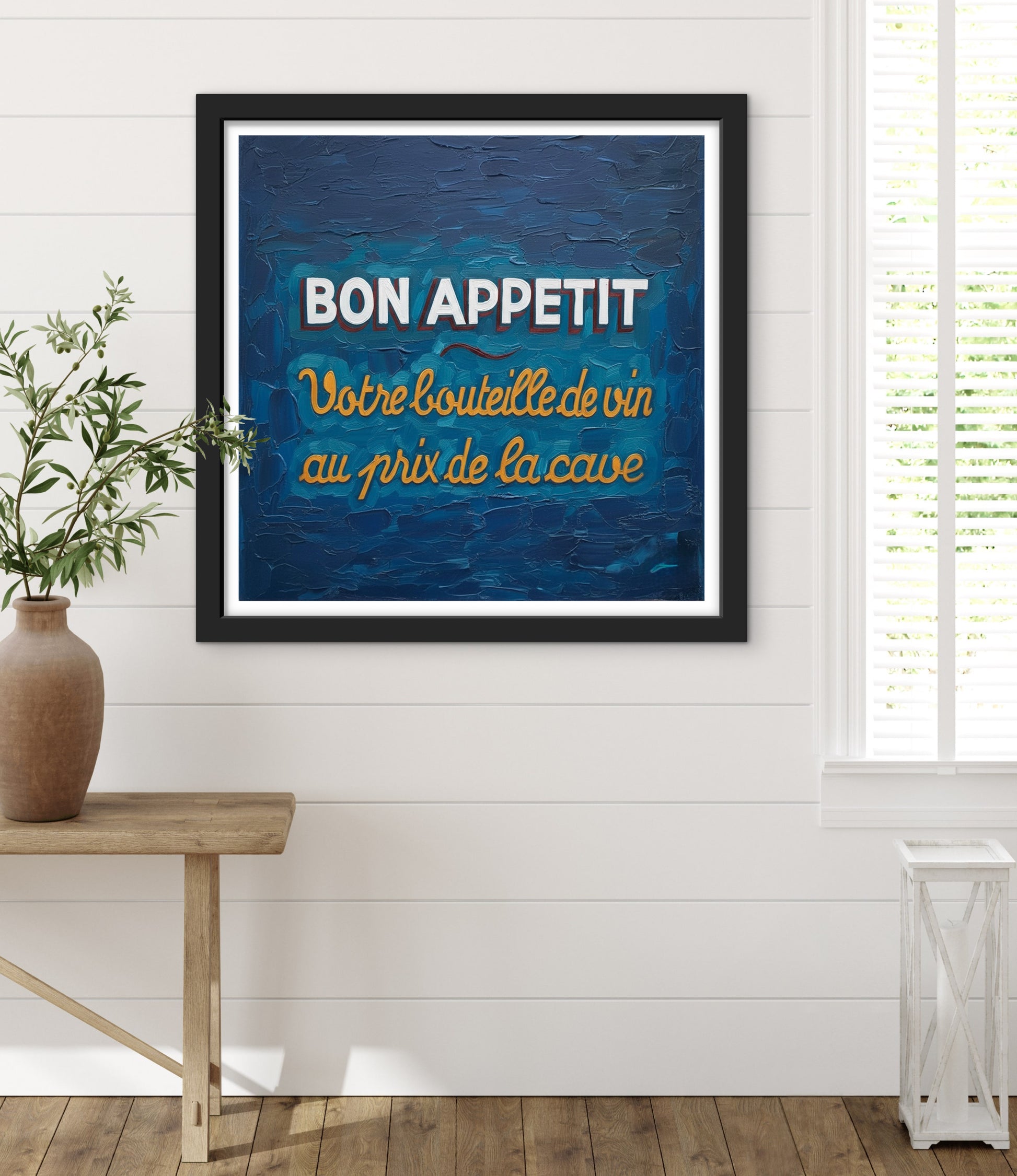 Framed artwork with 'Bon Appetit' text on a blue background in a room with a wooden table and plant.