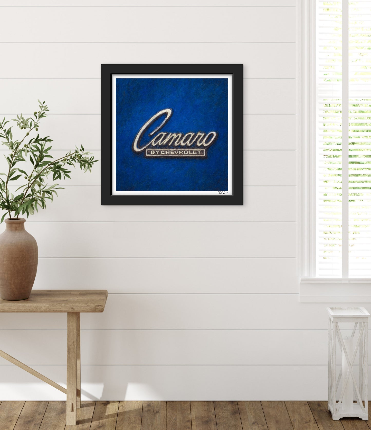 Framed artwork of the 'Camaro by Chevrolet' logo on a blue background in a room with a wooden table and plant.
