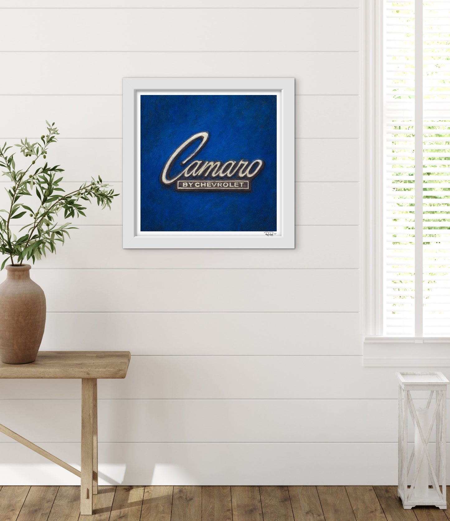Framed artwork with 'Camaro by Chevrolet' logo on a blue background, placed on a wooden table against a white wall.