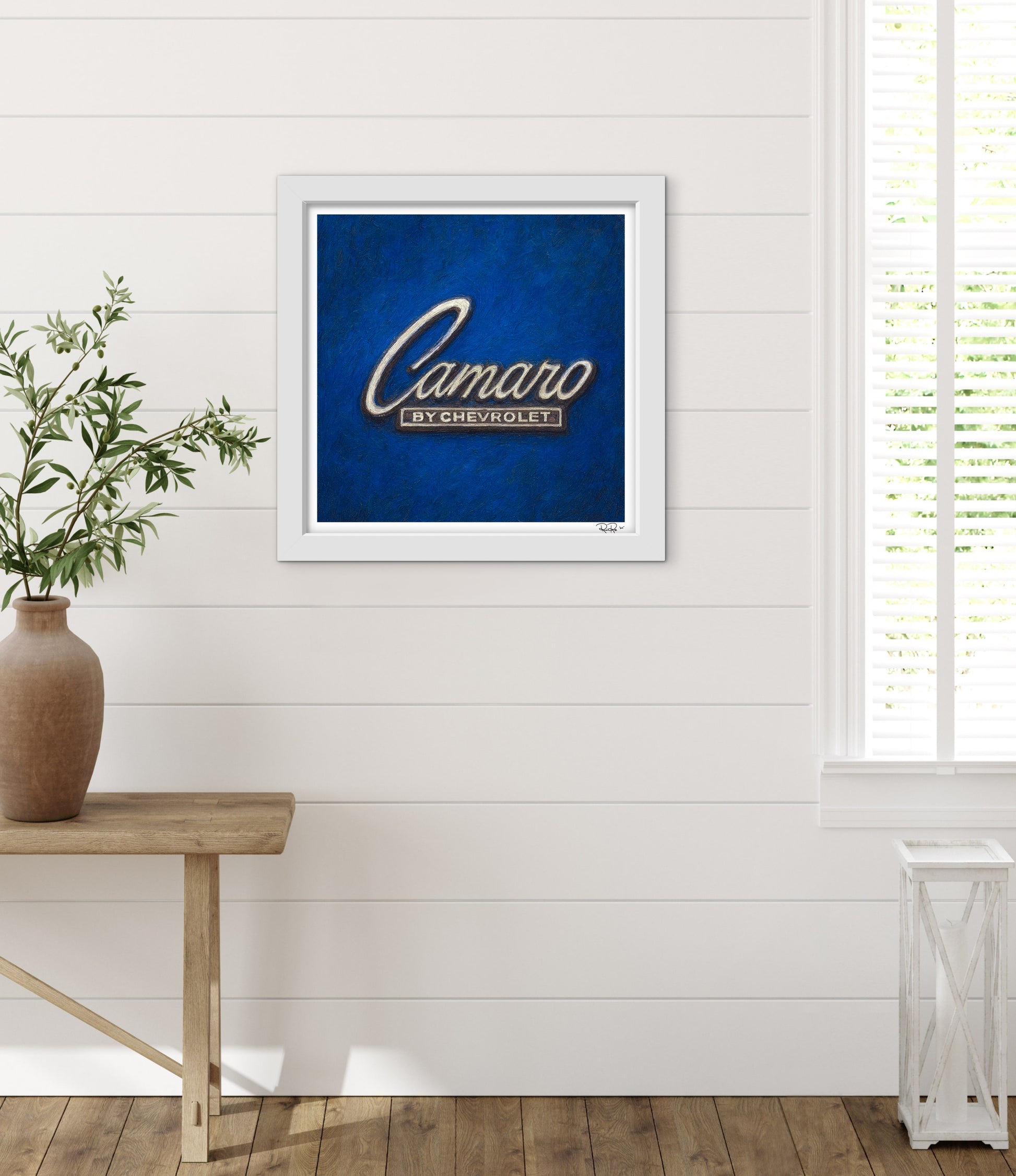 Framed artwork with 'Camaro by Chevrolet' logo on a blue background, placed on a wooden table against a white wall.