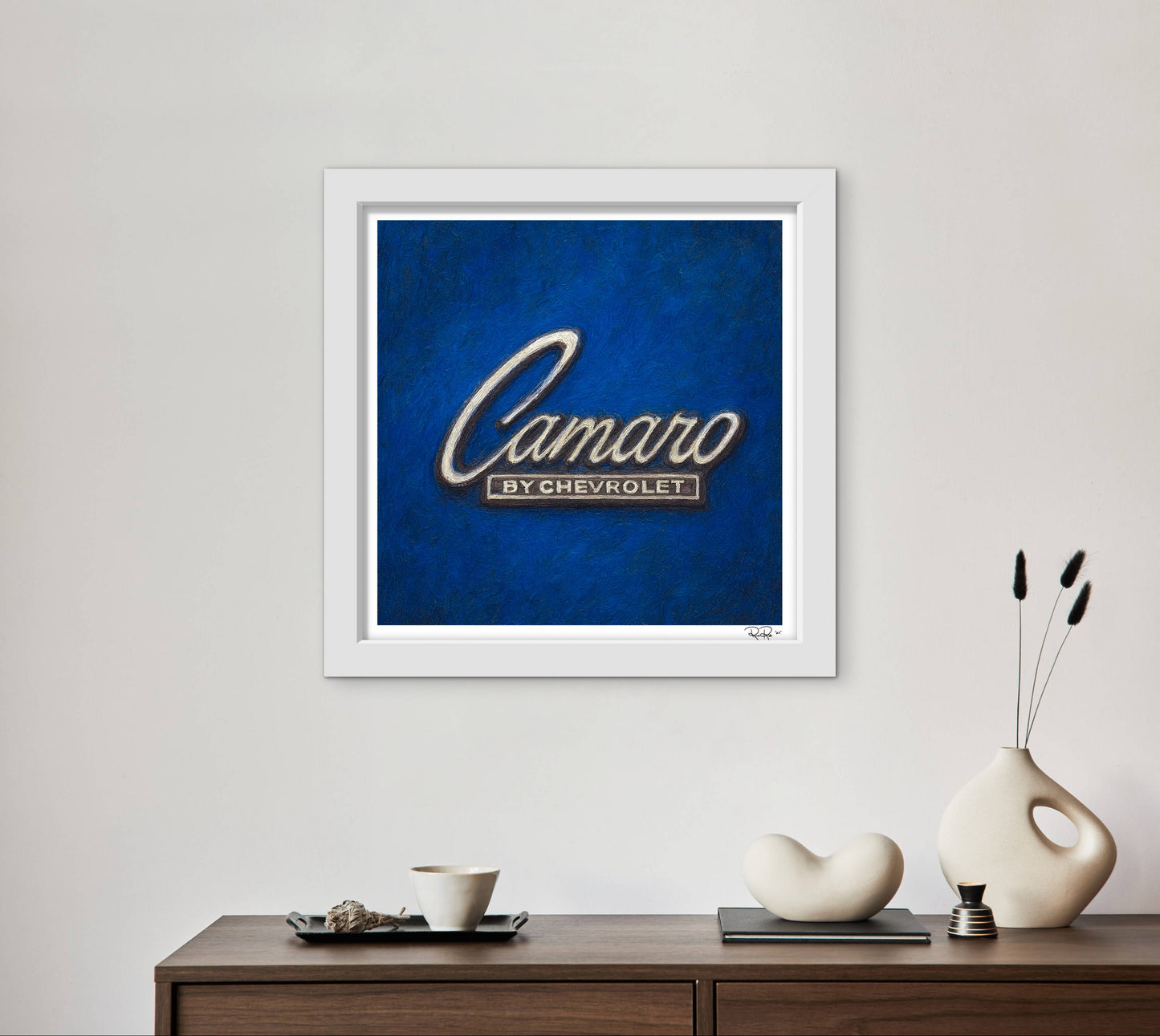 Framed artwork with 'Camaro by Chevrolet' on a blue background, displayed on a wall above a wooden console table.