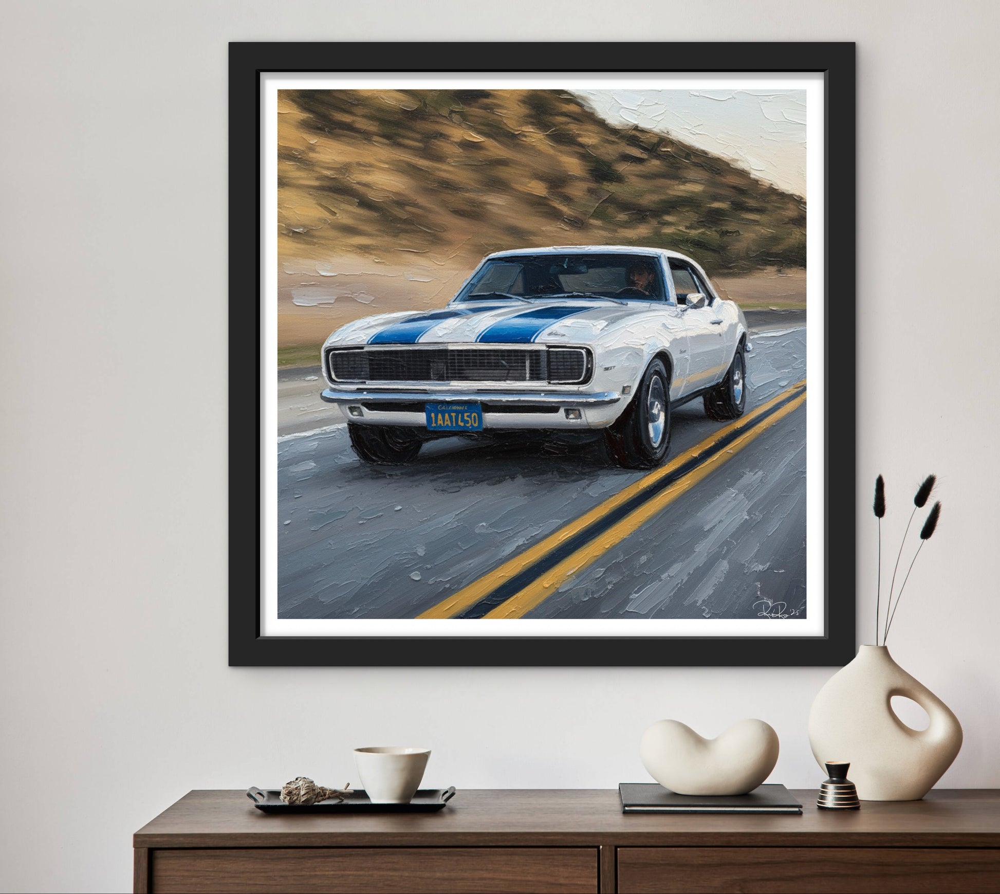 Framed artwork of a classic car on a road with mountains in the background, displayed on a wall above a wooden console table.
