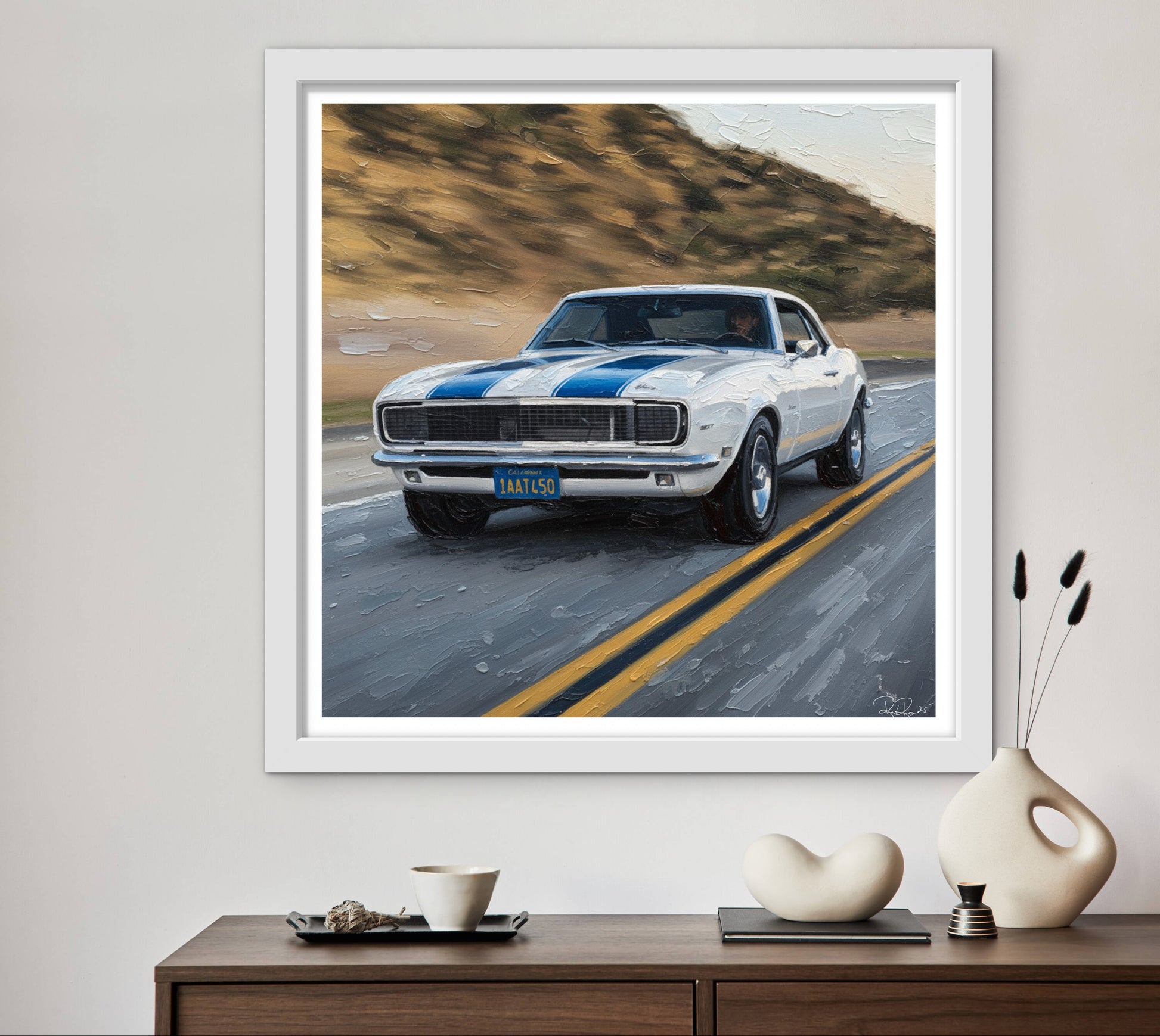 Framed artwork of a classic car on a road with mountains in the background, displayed on a wall above a wooden console table.