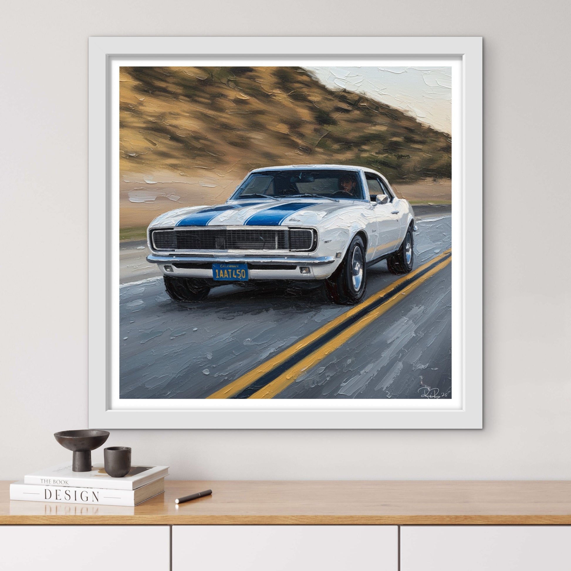 Framed artwork of a classic car on a road with mountains in the background, displayed on a wall above a wooden shelf.