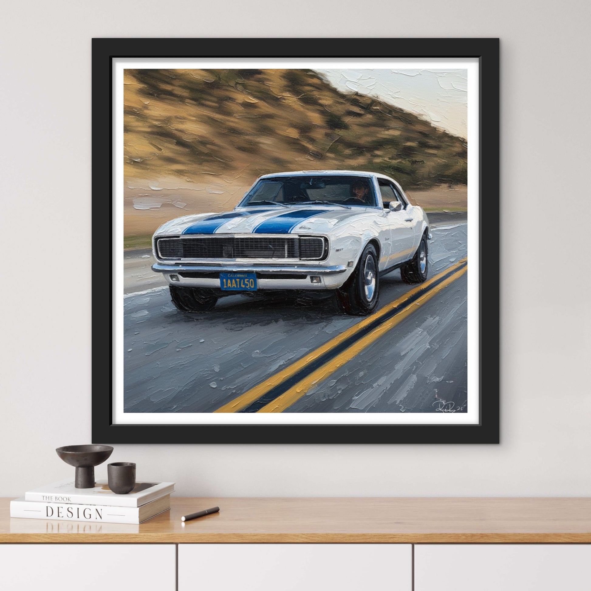 Framed artwork of a classic car on a road with mountains in the background, displayed on a wall above a wooden shelf.
