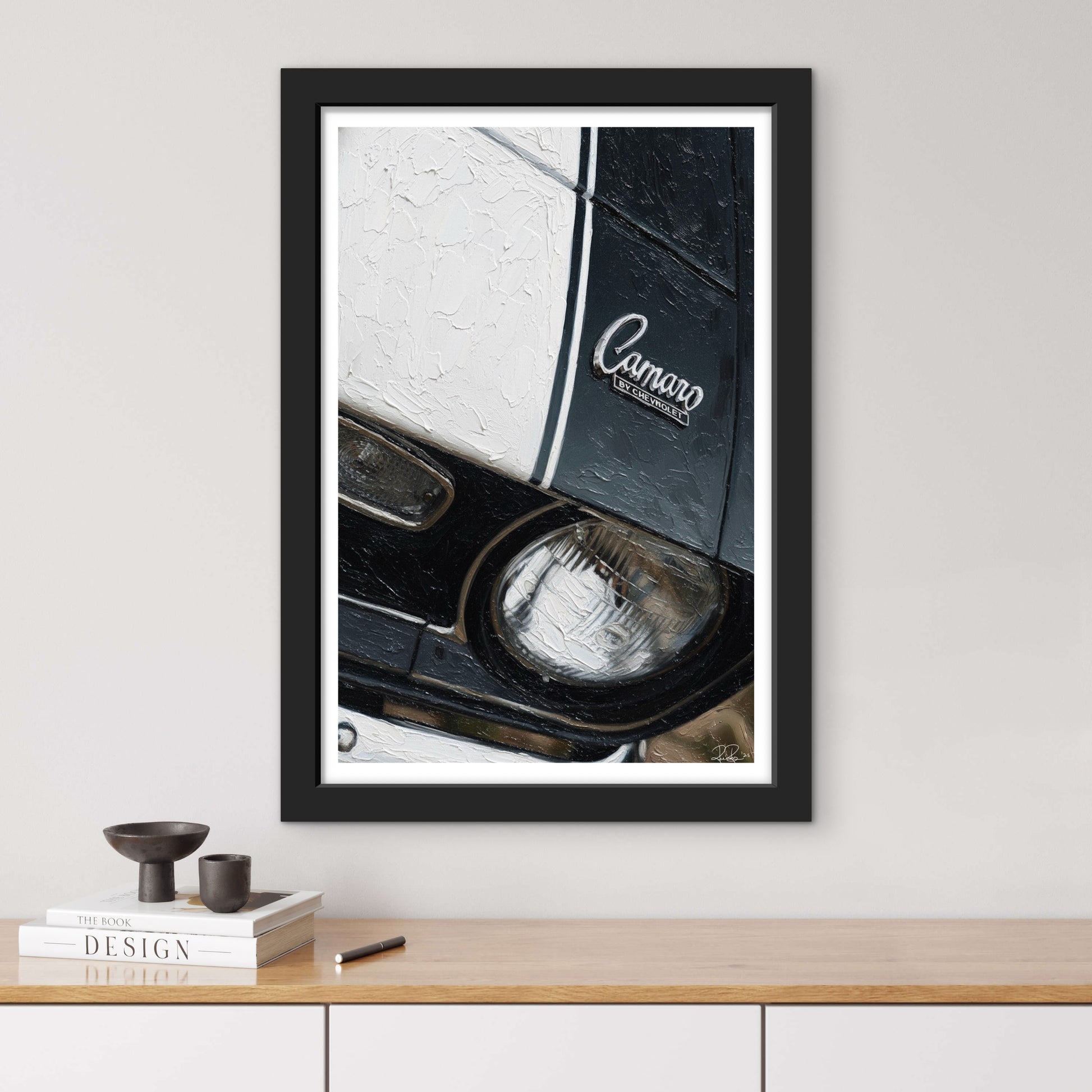 Framed artwork of a car headlight with 'Camaro' branding on a wall above a wooden shelf.