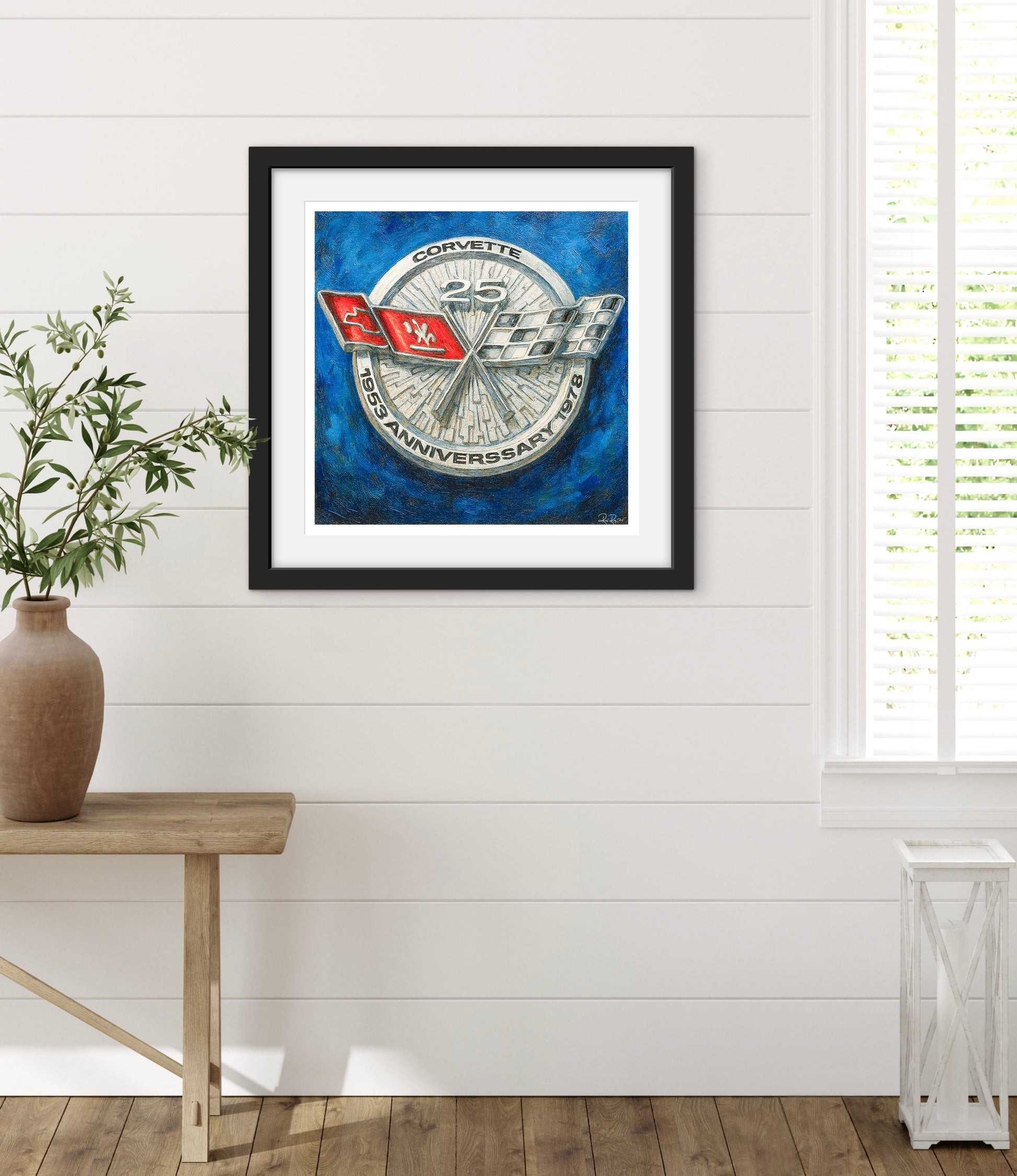 Framed Corvette logo art print on a white wall with a wooden table and plant.