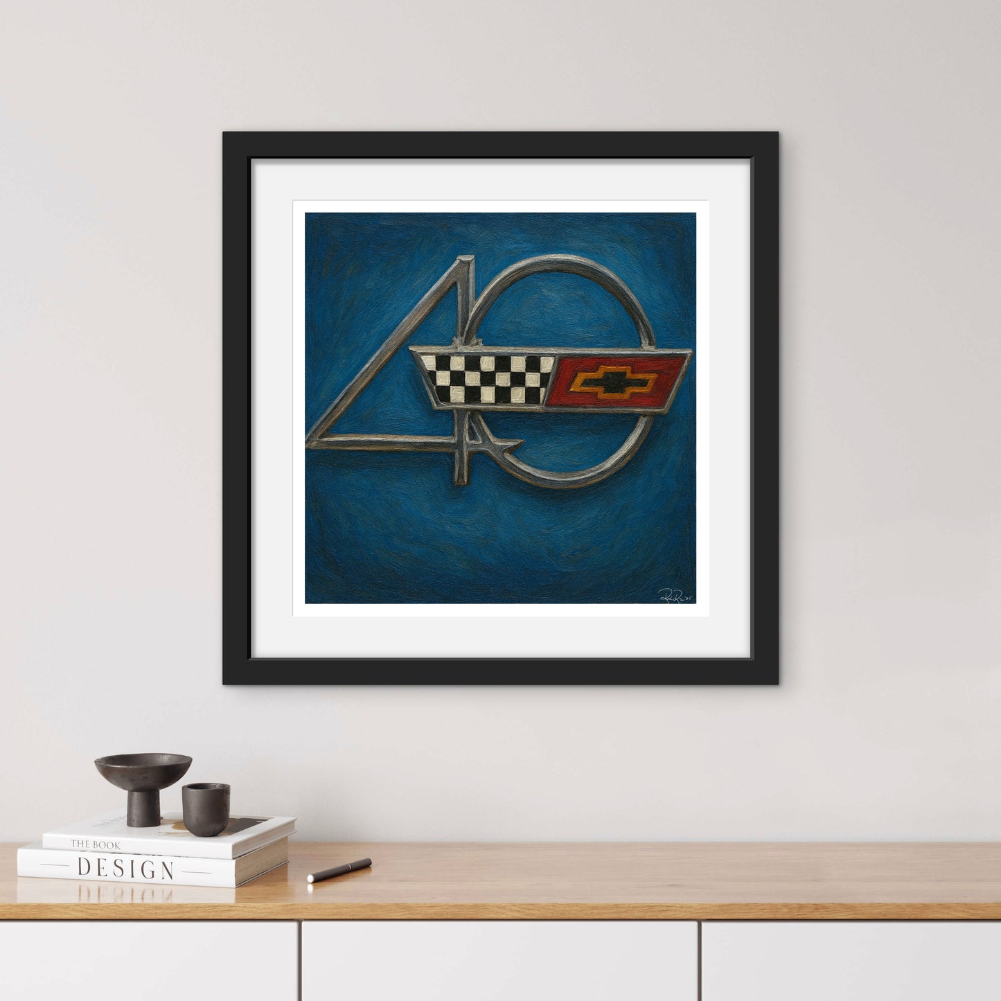 Framed artwork of a car emblem on a blue background, displayed on a wall above a wooden shelf.