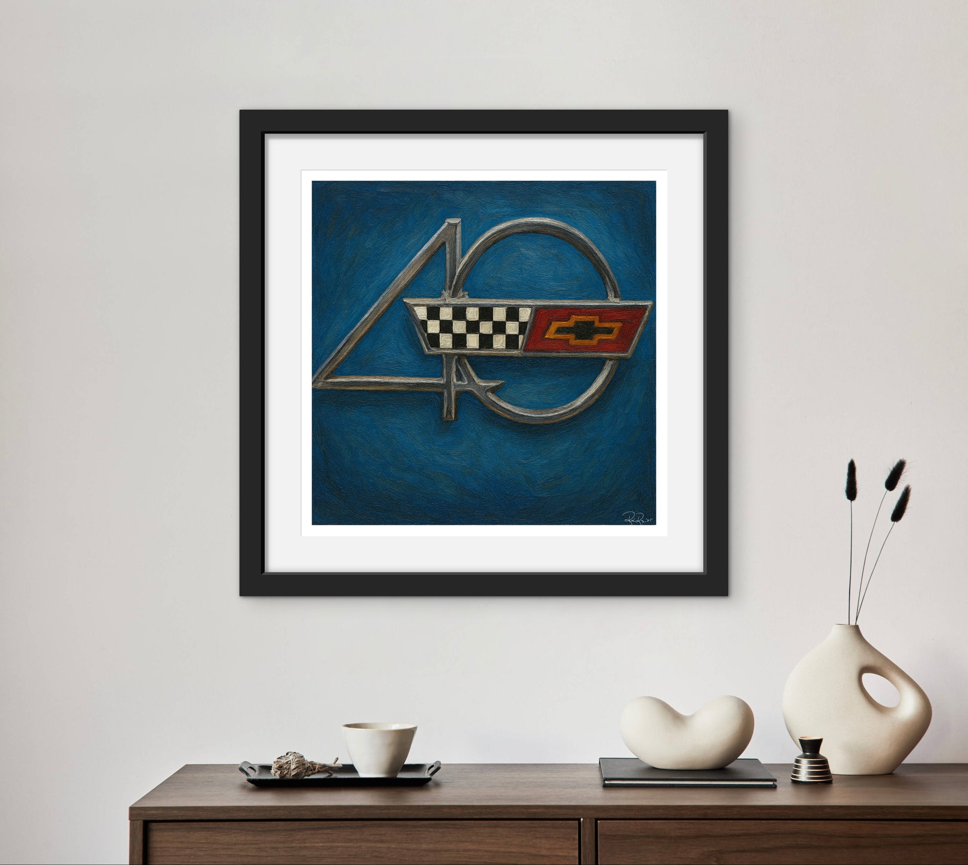Framed artwork of a car emblem on a blue background, displayed on a wall above a wooden console table with decorative items.