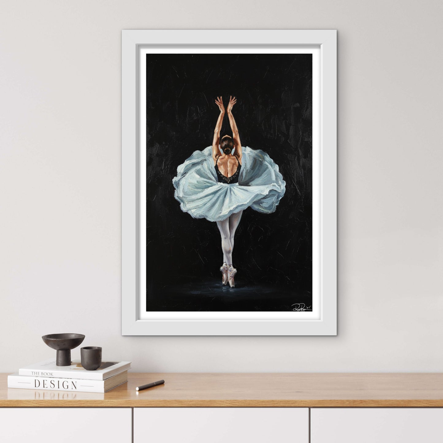 Framed artwork of a ballerina in a blue dress on a black background, displayed on a wall above a wooden shelf.