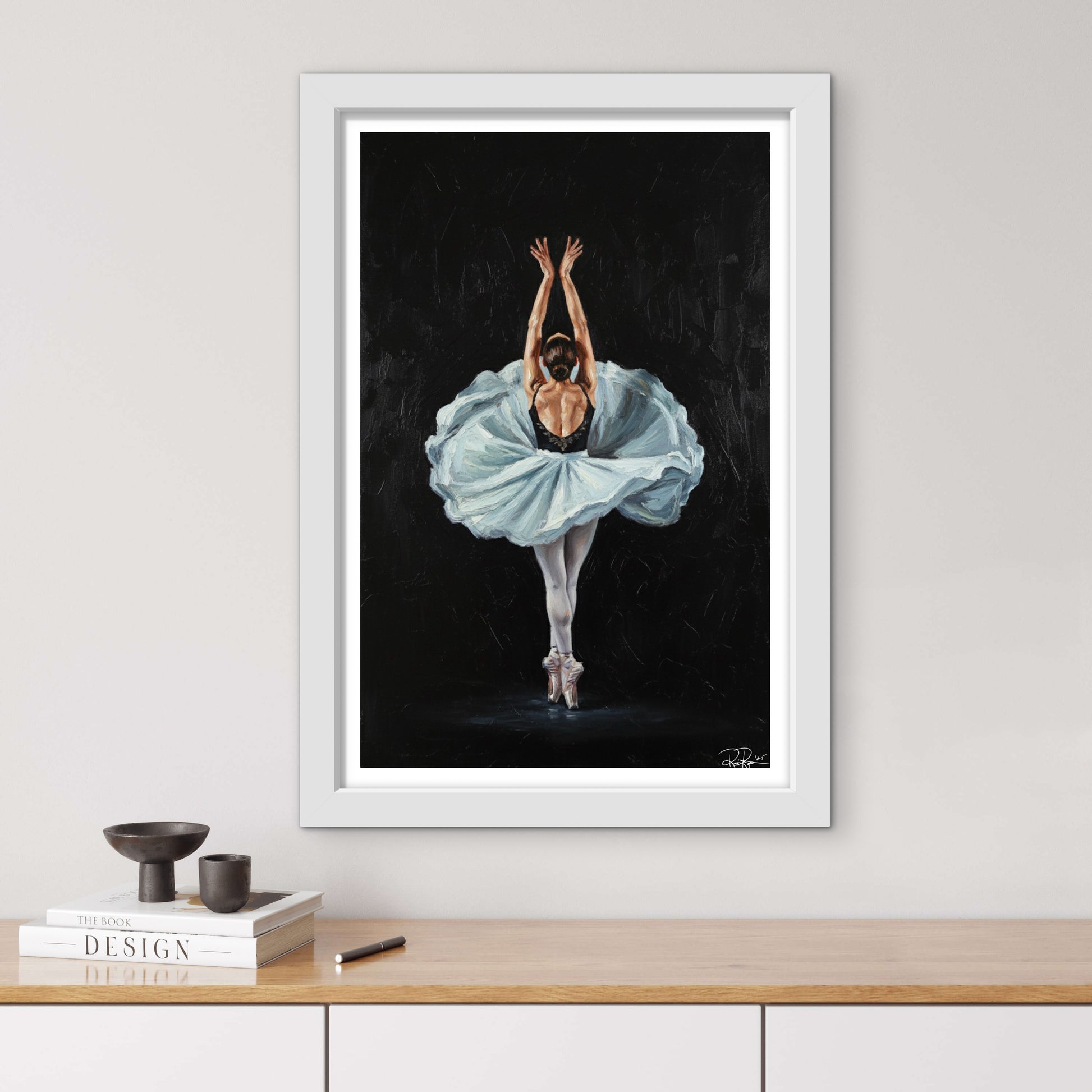 Framed artwork of a ballerina in a blue dress on a black background, displayed on a wall above a wooden shelf.