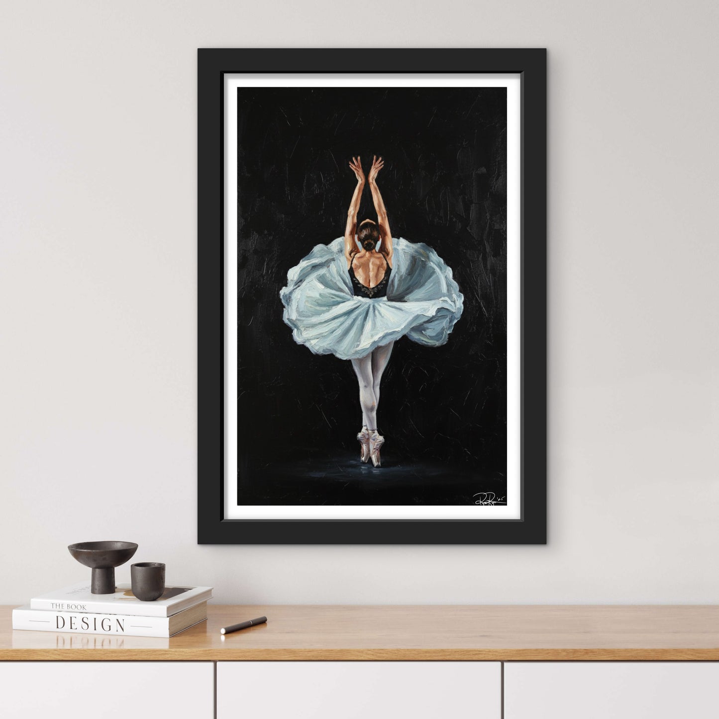Framed artwork of a ballerina in a blue dress on a black background, displayed on a wall above a wooden surface.