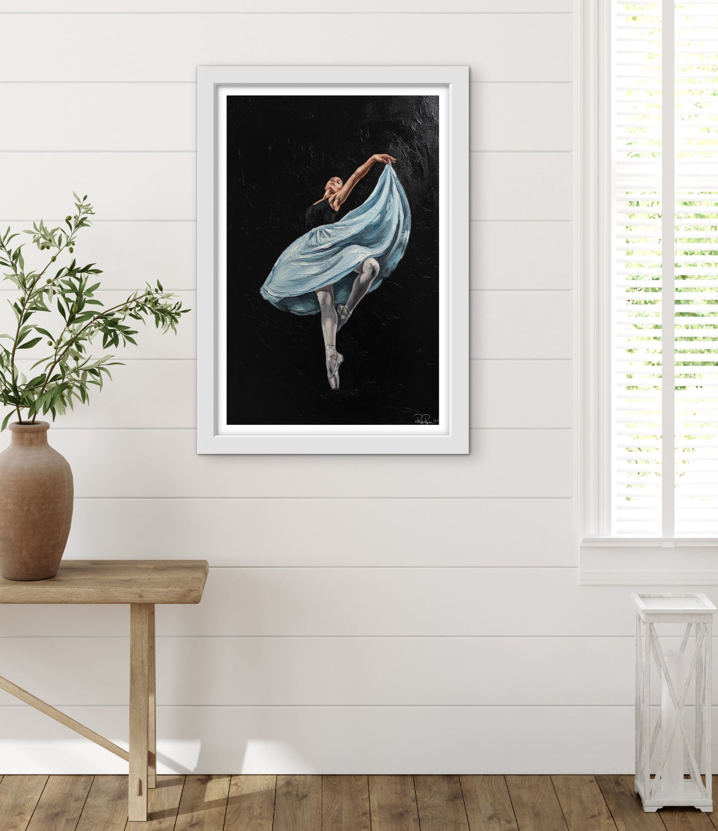 Framed artwork of a dancer in a blue dress on a white wall with a wooden table and plant.