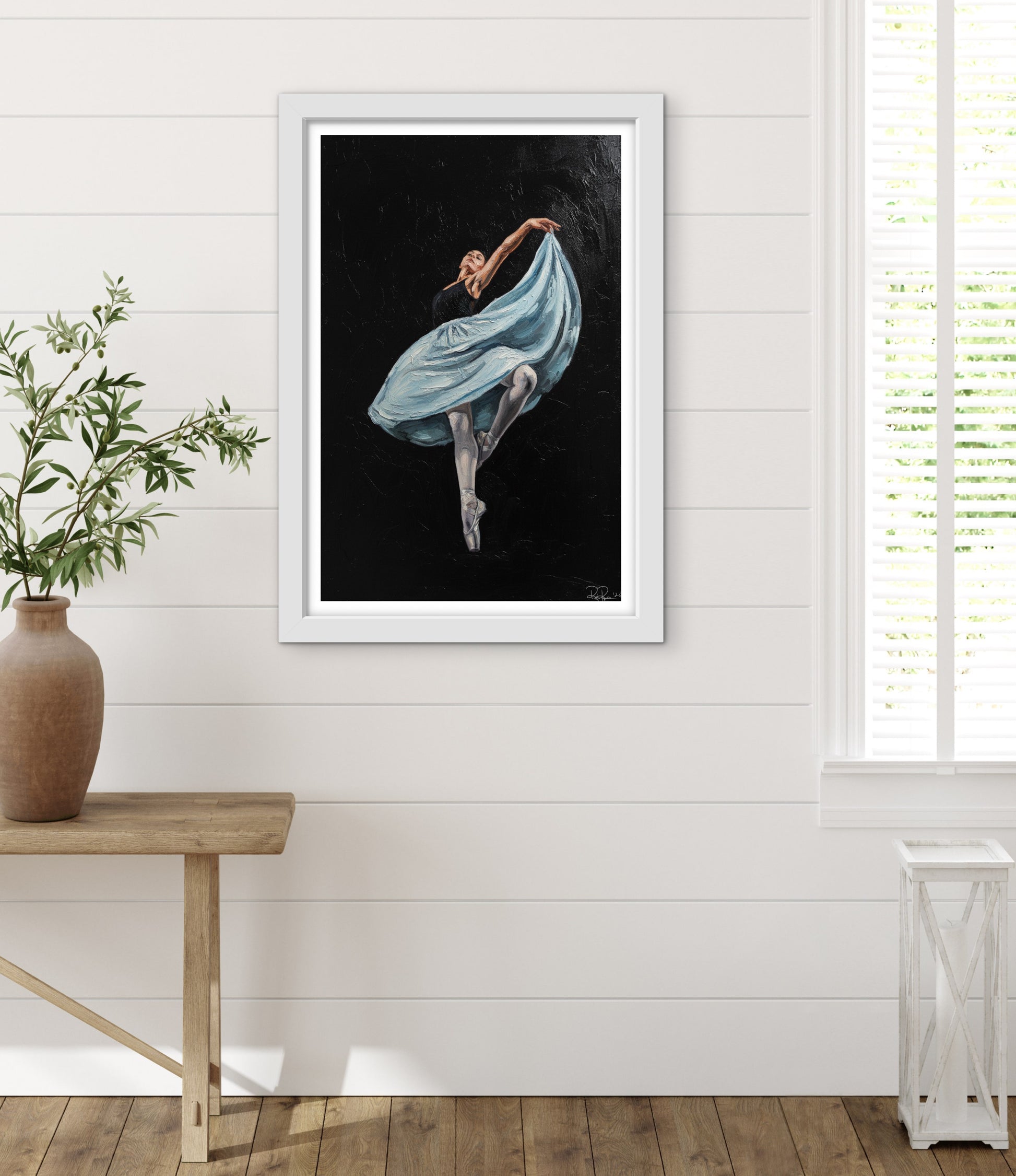 Framed artwork of a dancer in a blue dress on a white wall with a wooden table and plant.
