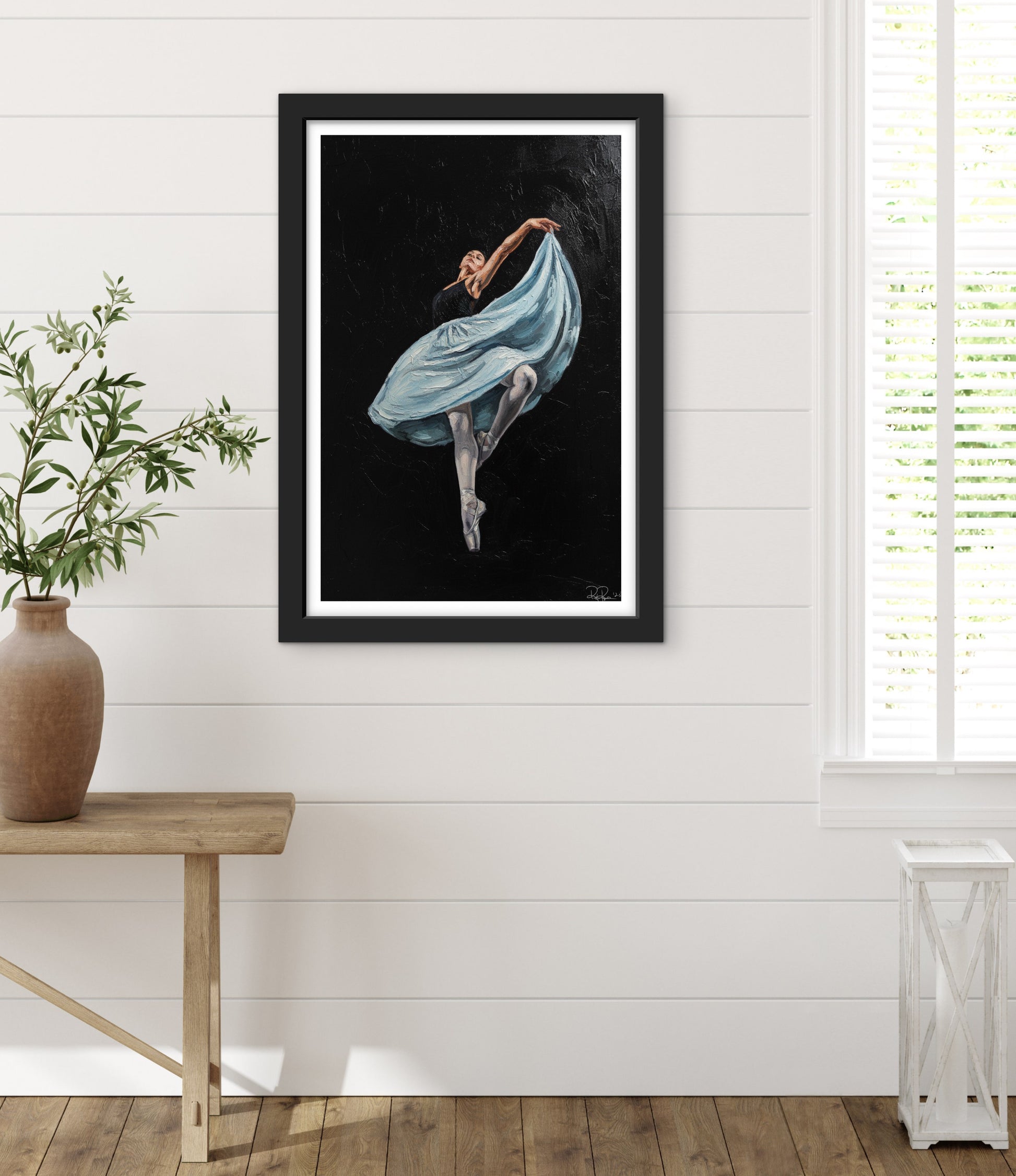 Framed artwork of a dancer in a blue dress on a white wall with a wooden table and plant.