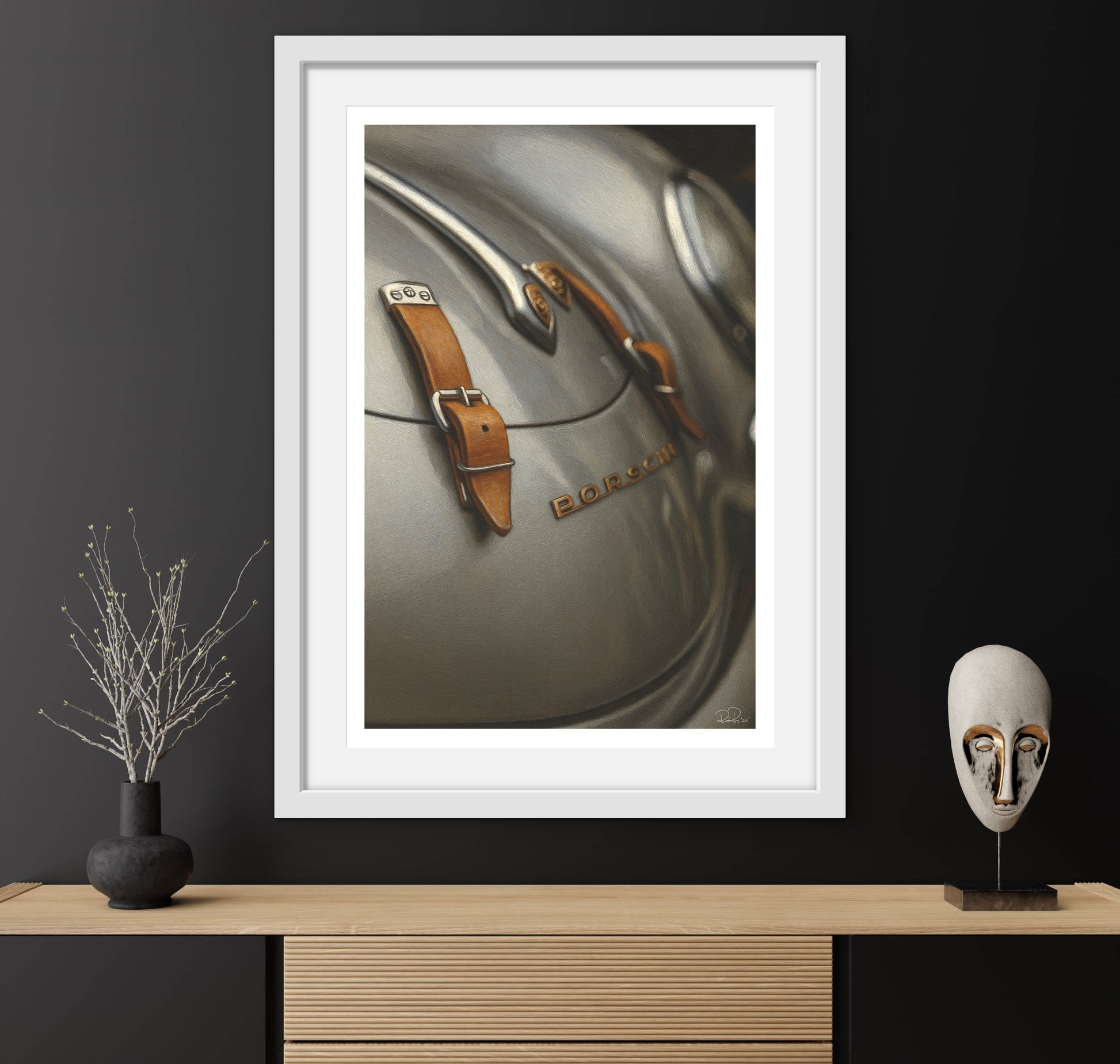 Framed artwork of a car emblem with a vase and mask on a wooden surface against a black wall.