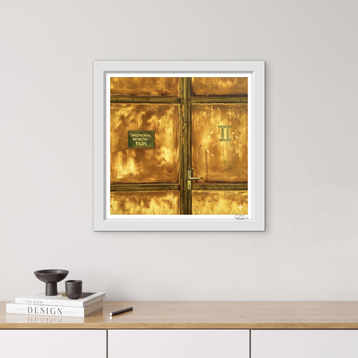 Framed artwork of a gold door on a white wall above a wooden surface with books and a pen.