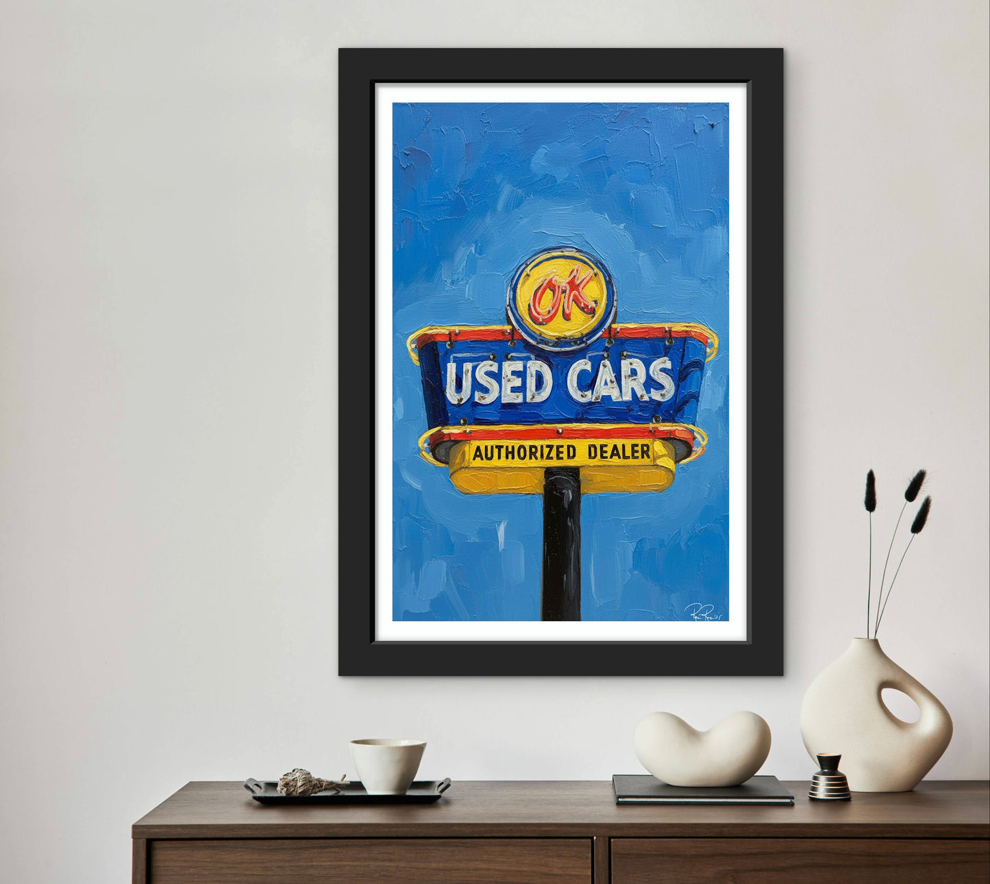 Framed artwork of a used car dealership sign on a wall above a wooden console table with decorative items.