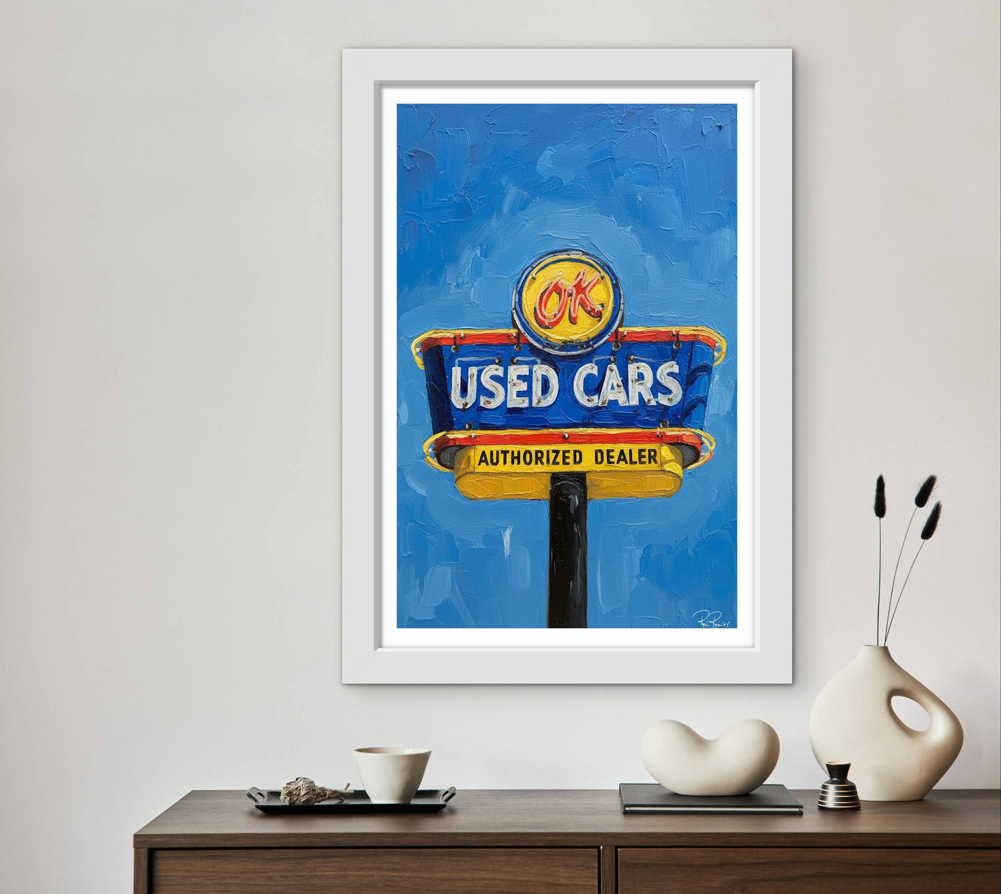 Framed artwork of a used car dealership sign on a wall above a wooden console table.