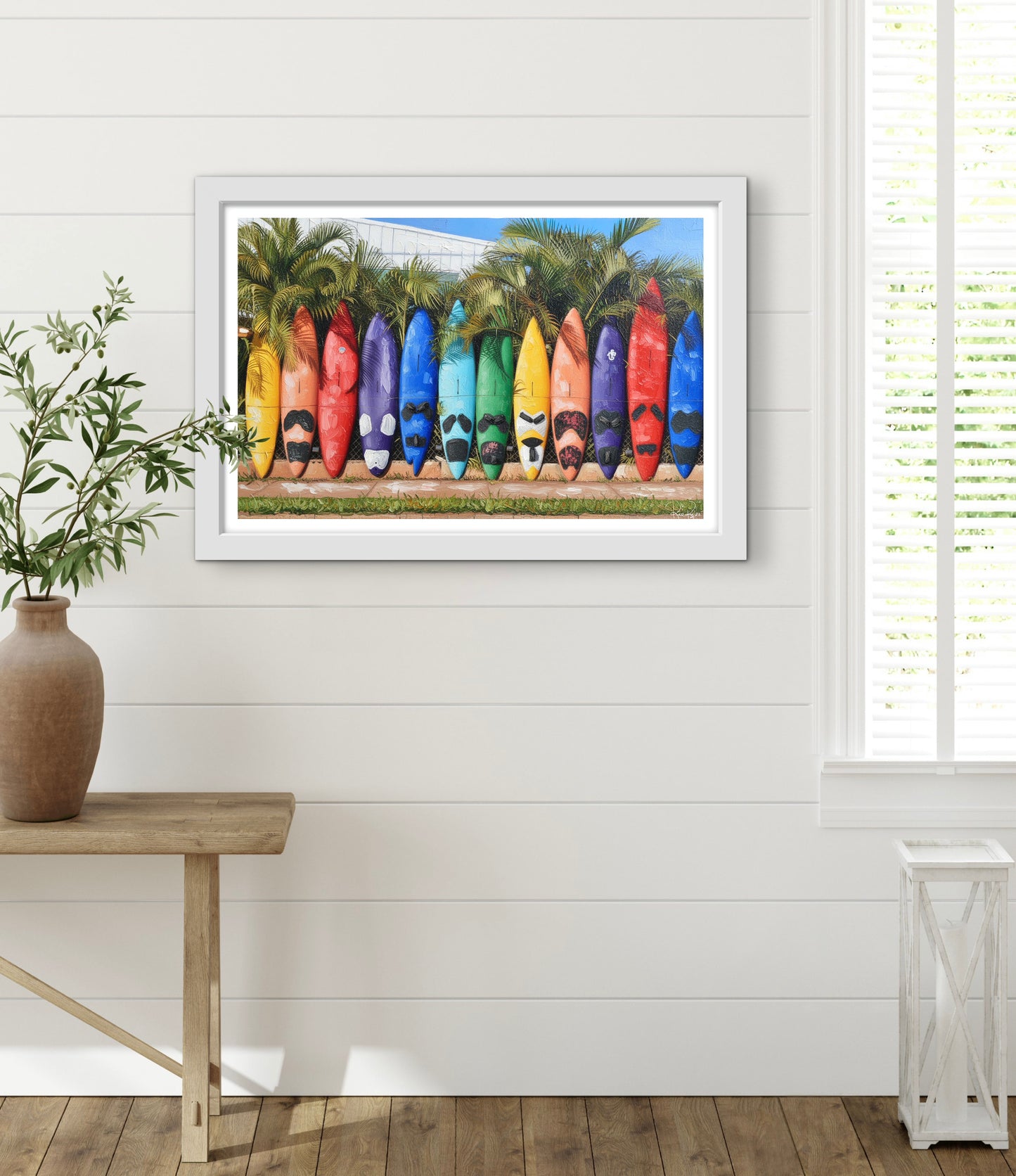 Framed artwork of colorful surfboards on a white wall with a wooden table and plant in the foreground.