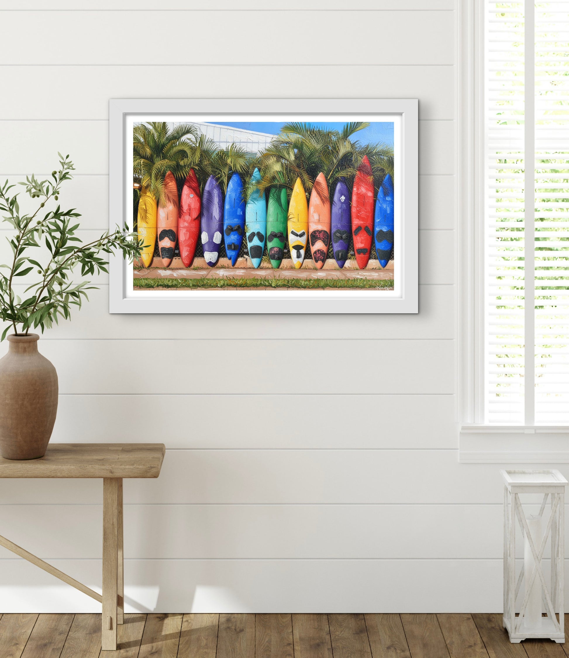 Framed artwork of colorful surfboards on a white wall with a wooden table and plant in the foreground.