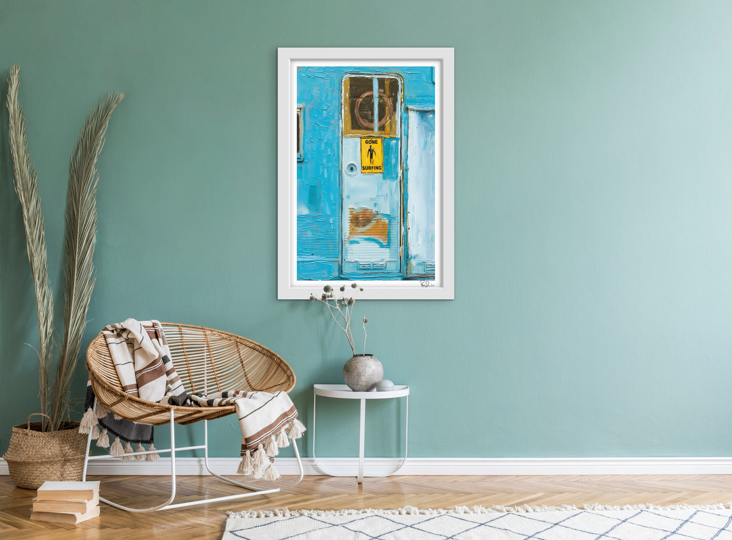 Framed artwork on a teal wall above a wicker chair with a blanket.