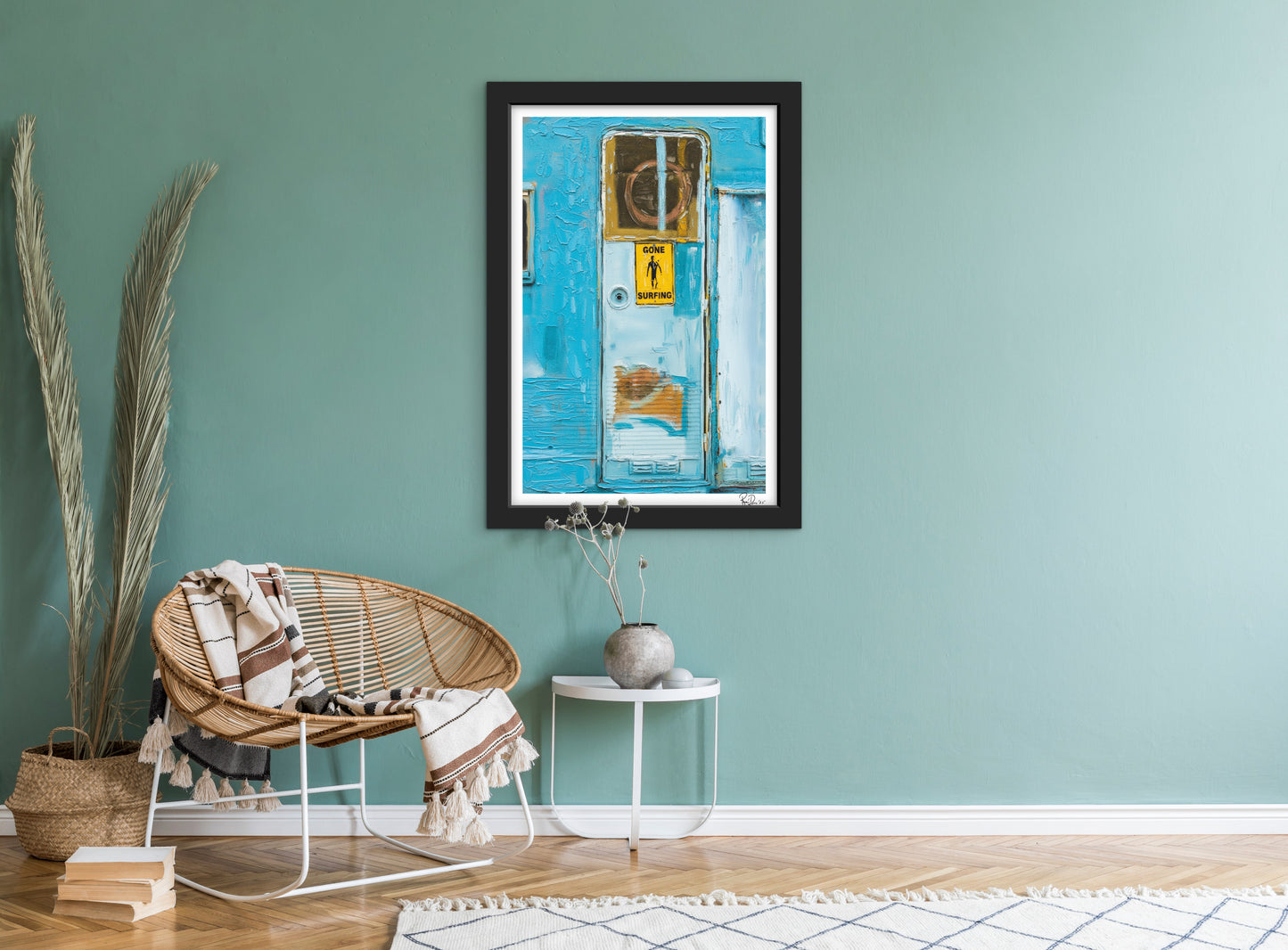Framed artwork on a teal wall above a wicker chair with a blanket.