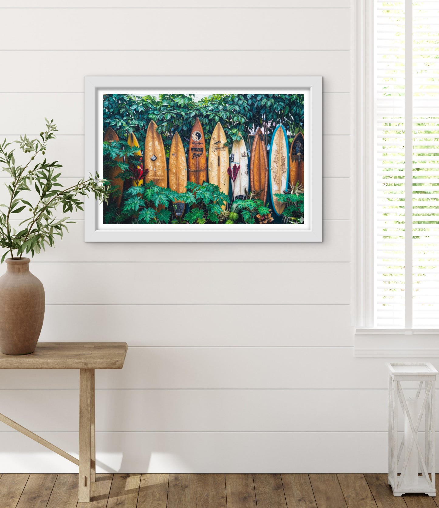 Framed artwork of surfboards in a tropical setting on a white wall with a wooden table and plant.