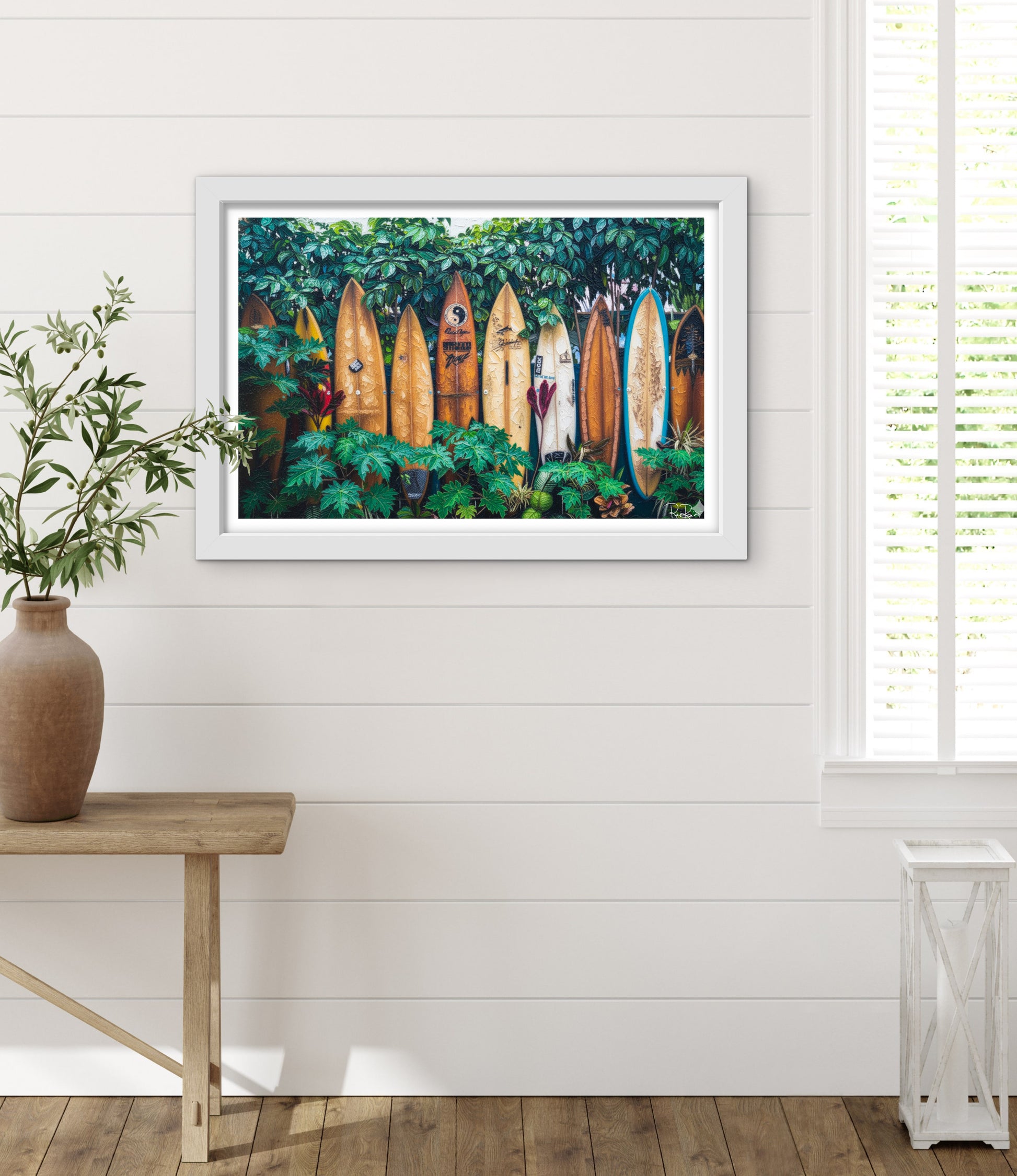 Framed artwork of surfboards in a tropical setting on a white wall with a wooden table and plant.