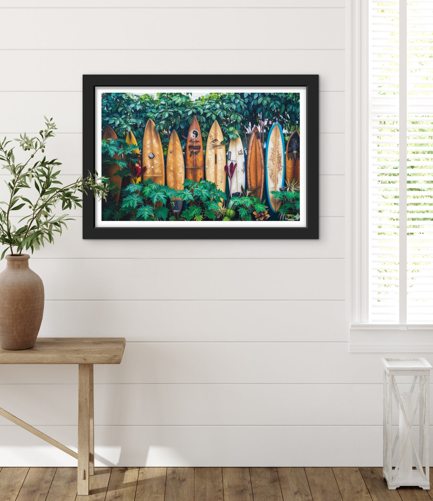 Framed artwork of surfboards in a forest displayed on a wall with a plant and table in the foreground.