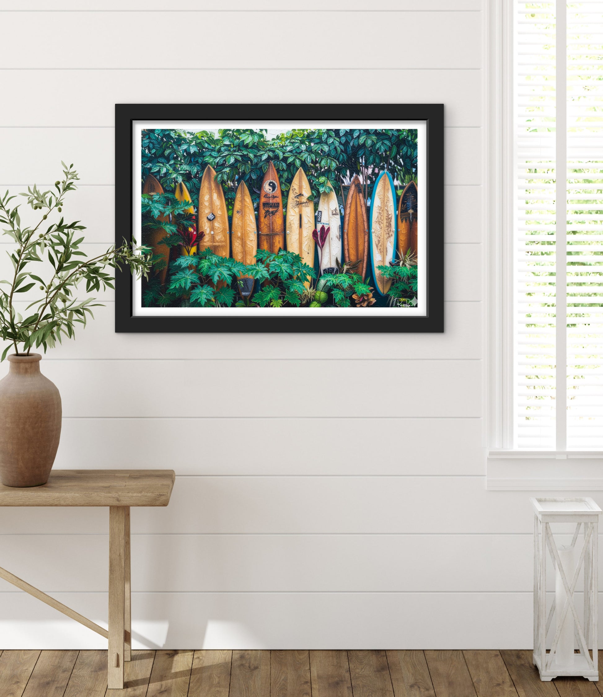 Framed artwork of surfboards in a forest displayed on a wall with a plant and table in the foreground.